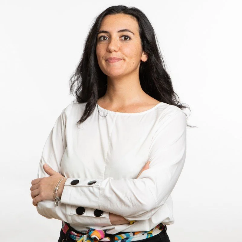 Una donna con capelli castani e vestito bianco, con le braccia incrociate, sfondo bianco.