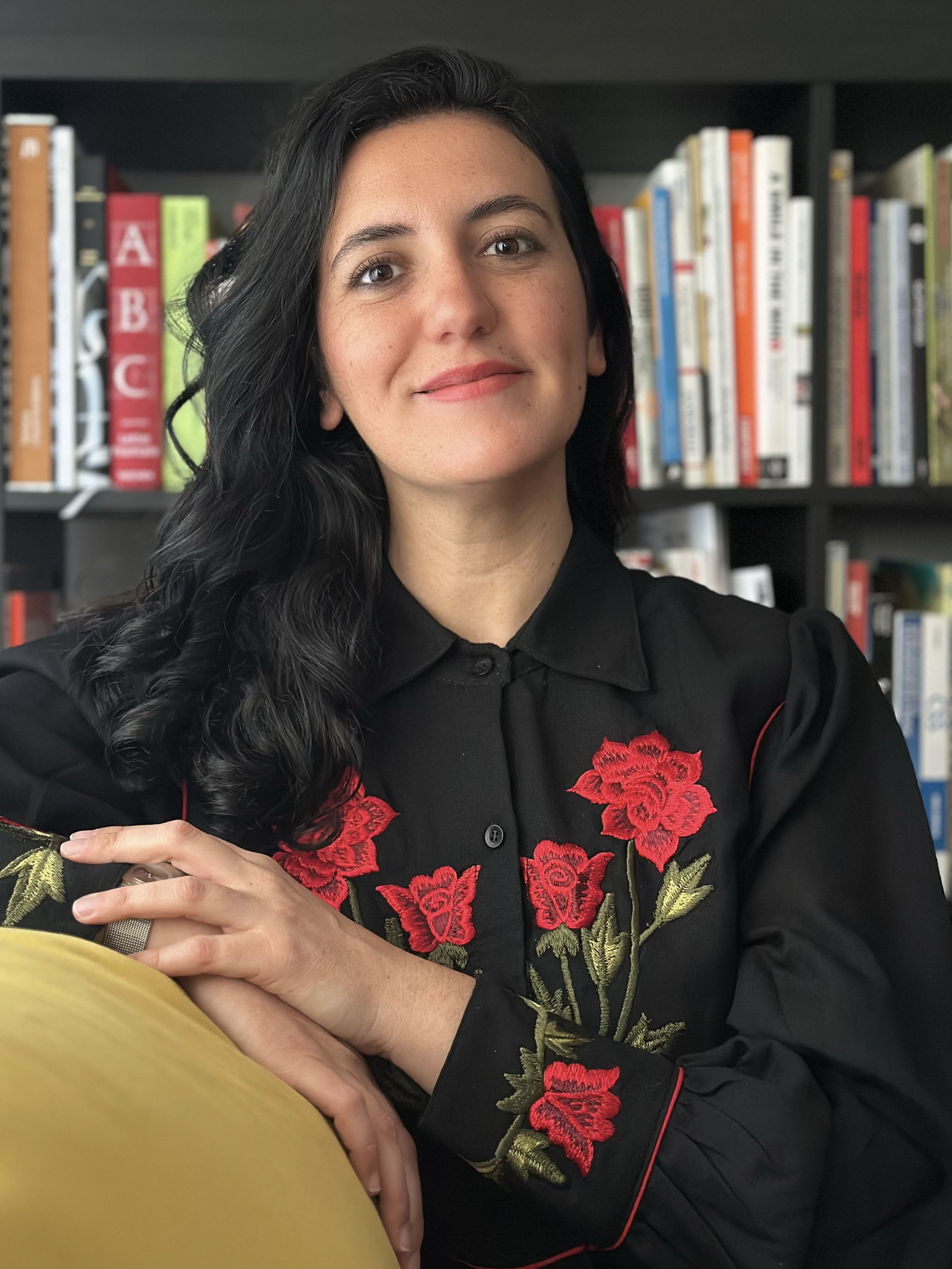 Donna con capelli neri ricci, vestita con camicia nera decorata con ricami di fiori rossi e foglie verdi, seduta di fronte a una libreria con vari libri colorati.