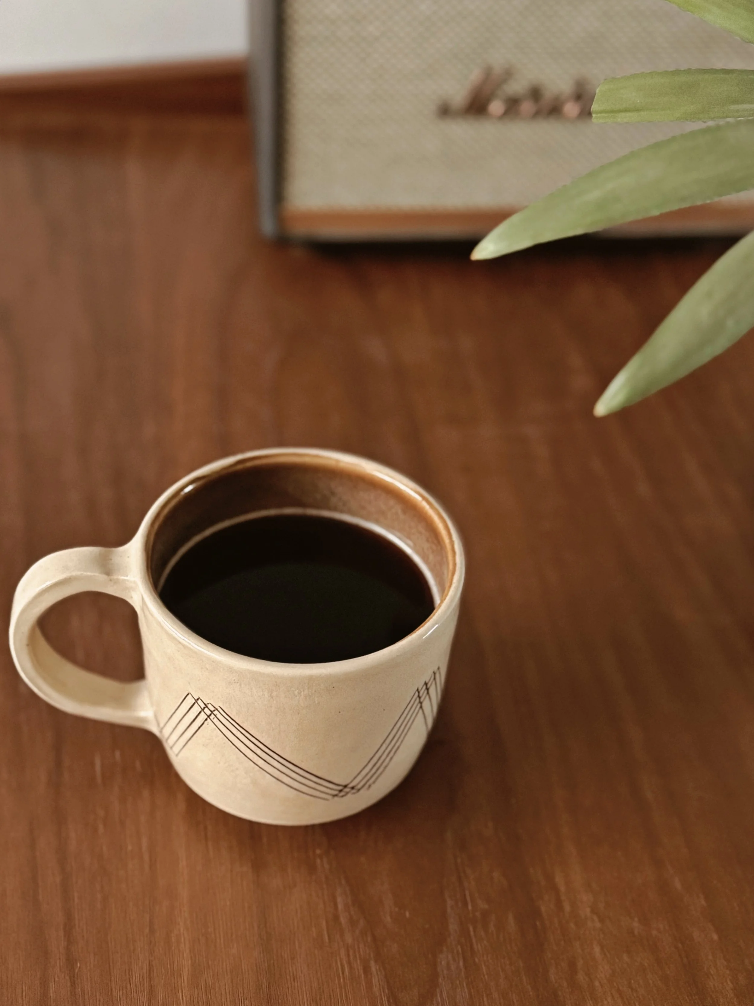 A white ceramic coffee mug with a black geometric line pattern on a wooden table, filled with black coffee, next to a green plant leaf and a framed picture in the background.