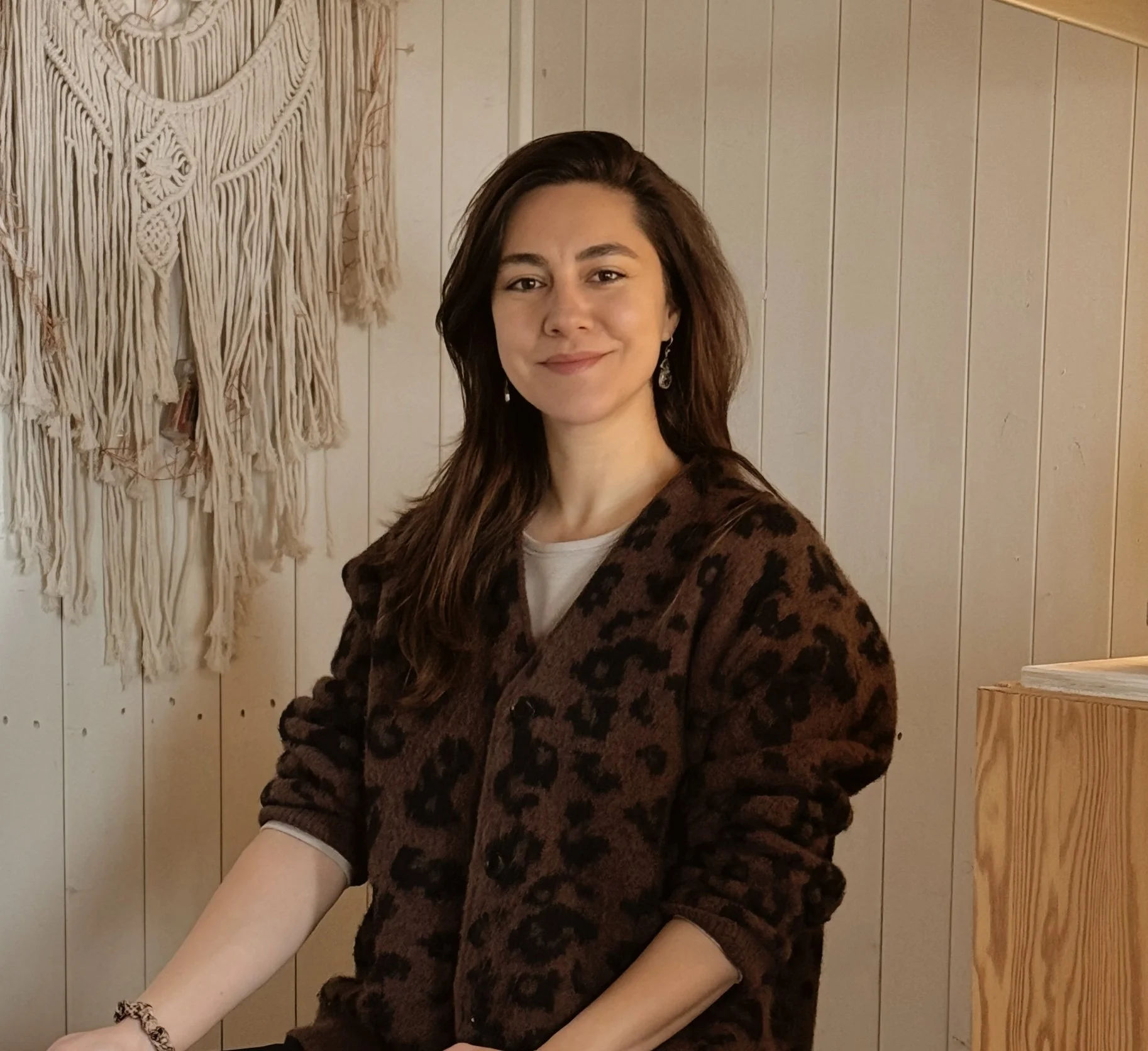 A woman with long brown hair smiling and wearing a leopard print cardigan, standing in front of a wooden wall with a large woven wall hanging.