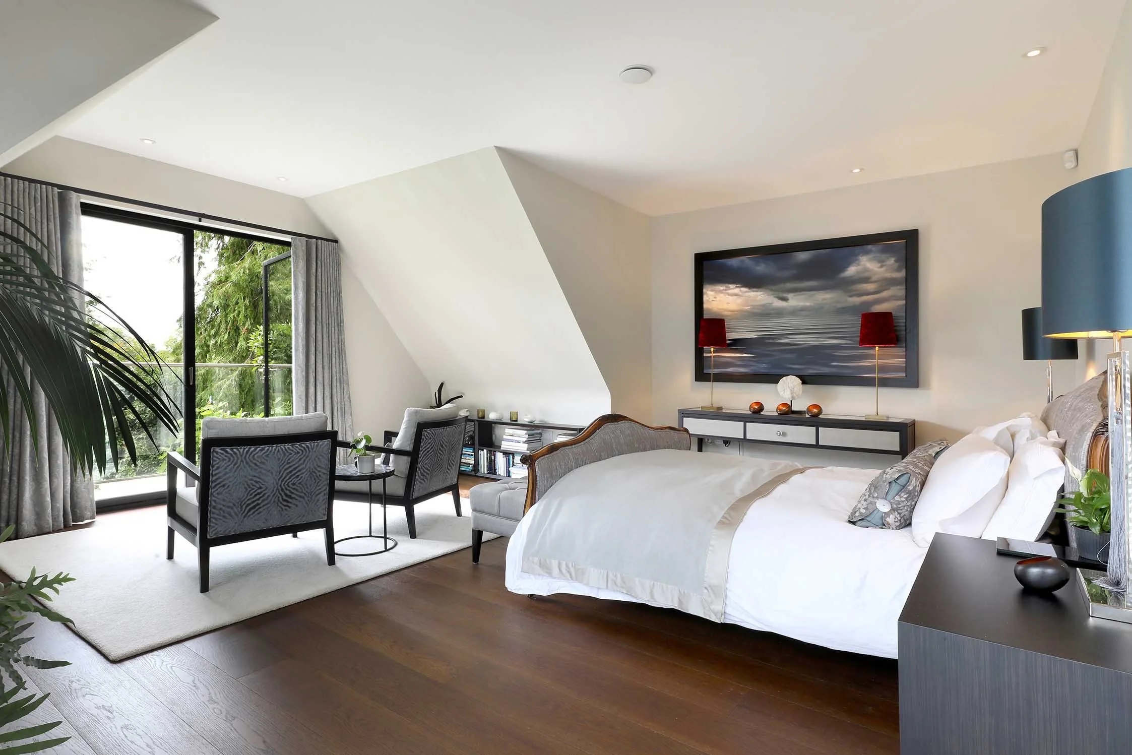 Modern bedroom with a large bed, a flat-screen TV on the wall, a sitting area with two chairs and a small table, a sliding glass door with curtains, and a view of greenery outside.