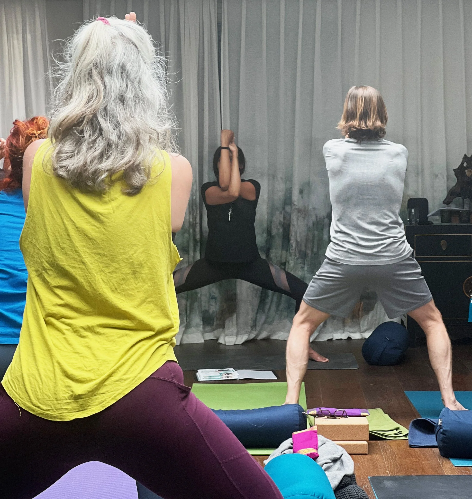Group of people in a yoga class, with an instructor demonstrating a pose in front of a grey curtain, some participants in a squat pose, and yoga mats on the floor.