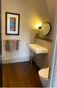 A bathroom with a wall-mounted sink, a round mirror, a yellow wall sconce, and a framed colorful artwork on the wall. There is a grey towel hanging on a rack and a white toilet in the foreground. The floor is wooden.