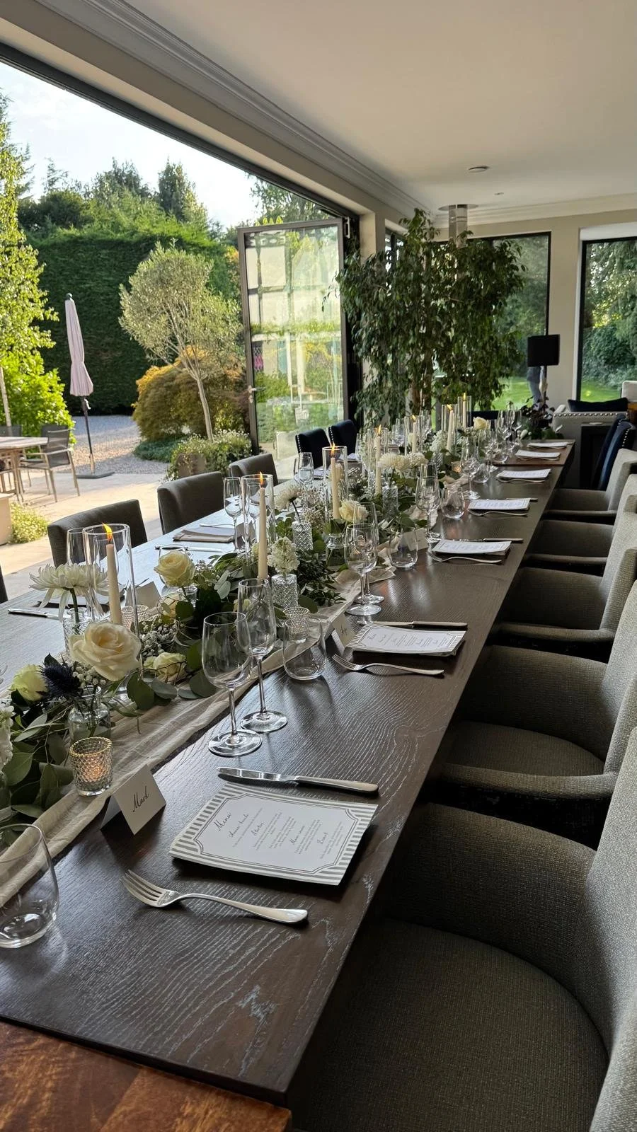 Elegantly set dining table with white floral arrangements, candles, and glassware in a bright room with large windows overlooking a garden