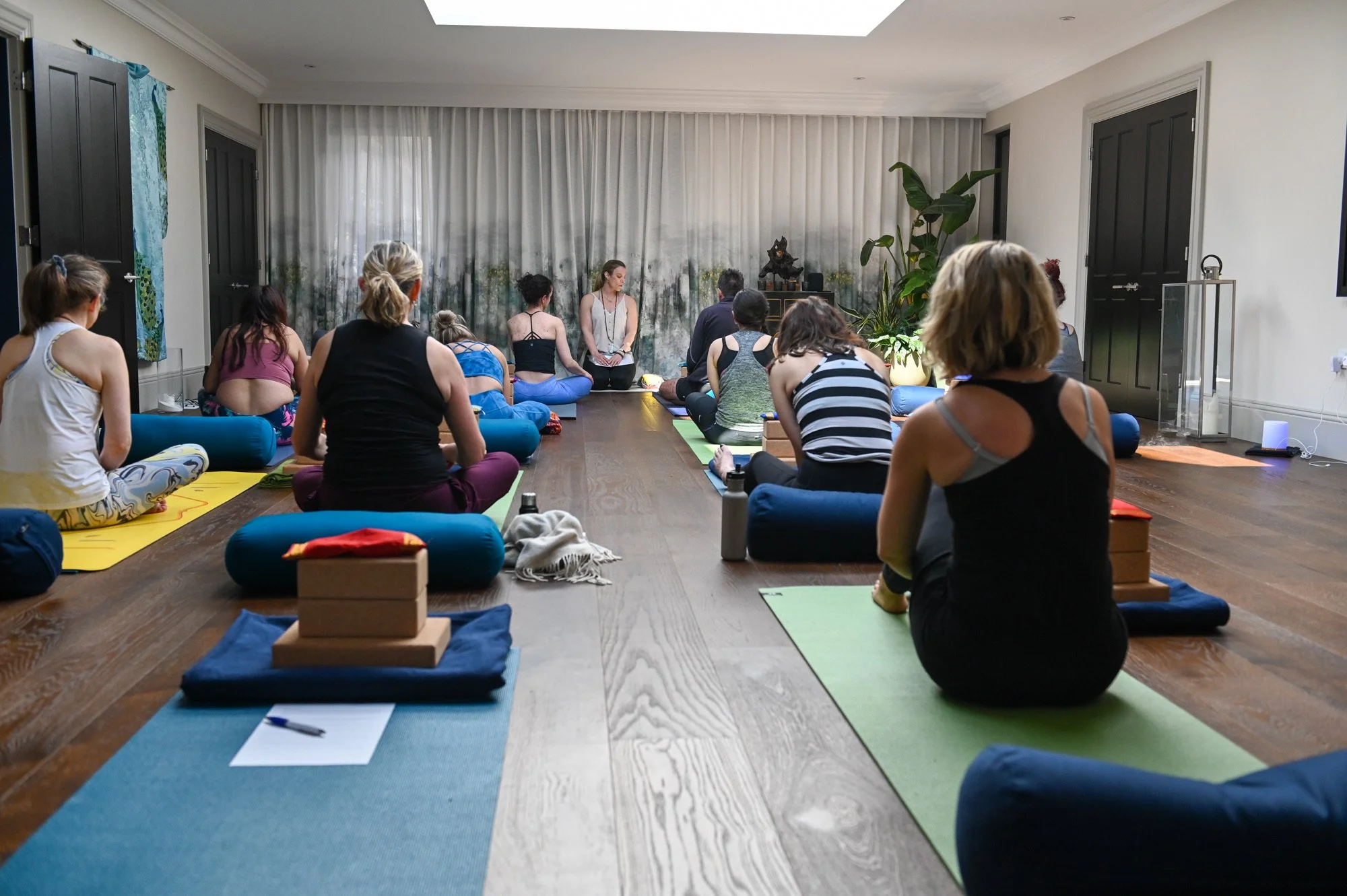 A group of people participating in a yoga class in a spacious indoor studio. They are seated on yoga mats facing a instructor at the front near a curtain and plant. Yoga props and water bottles are visible.