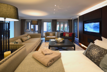 A modern hotel suite with a seating area, large windows, and a bed with a towel and pillow.