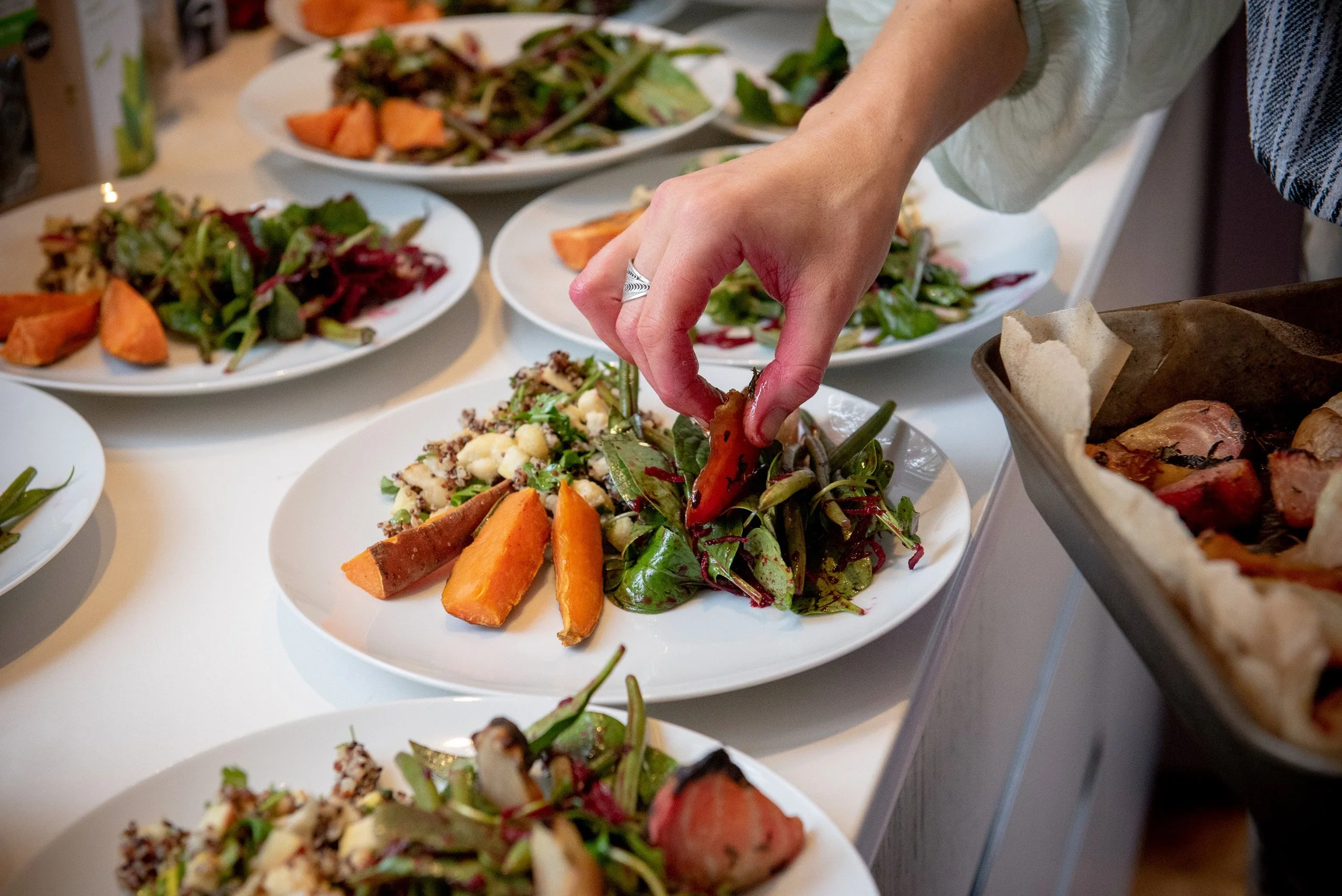 Person arranging a fresh green salad with sweet potatoes on a white plate, surrounded by other plates of salads and dishes on a table.