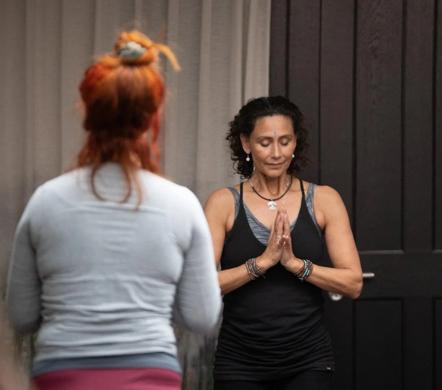 Two women in a meditative pose, with one woman facing away and the other in front with hands pressed together and eyes closed.