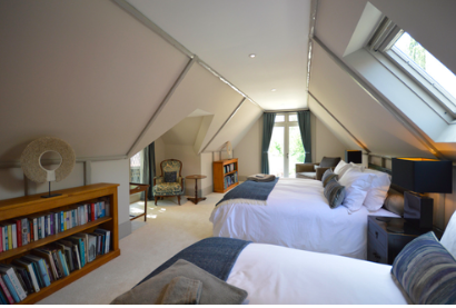 Attic bedroom with two beds, a bookshelf, a vintage armchair, and large windows.