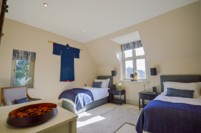 A bedroom with two beds, each with blue bedspreads and white pillows, a small black nightstand between them, and a window with a view outside. A traditional blue garment hangs on the wall, and a wooden chair is placed near the wall. Sunlight is coming through the window.