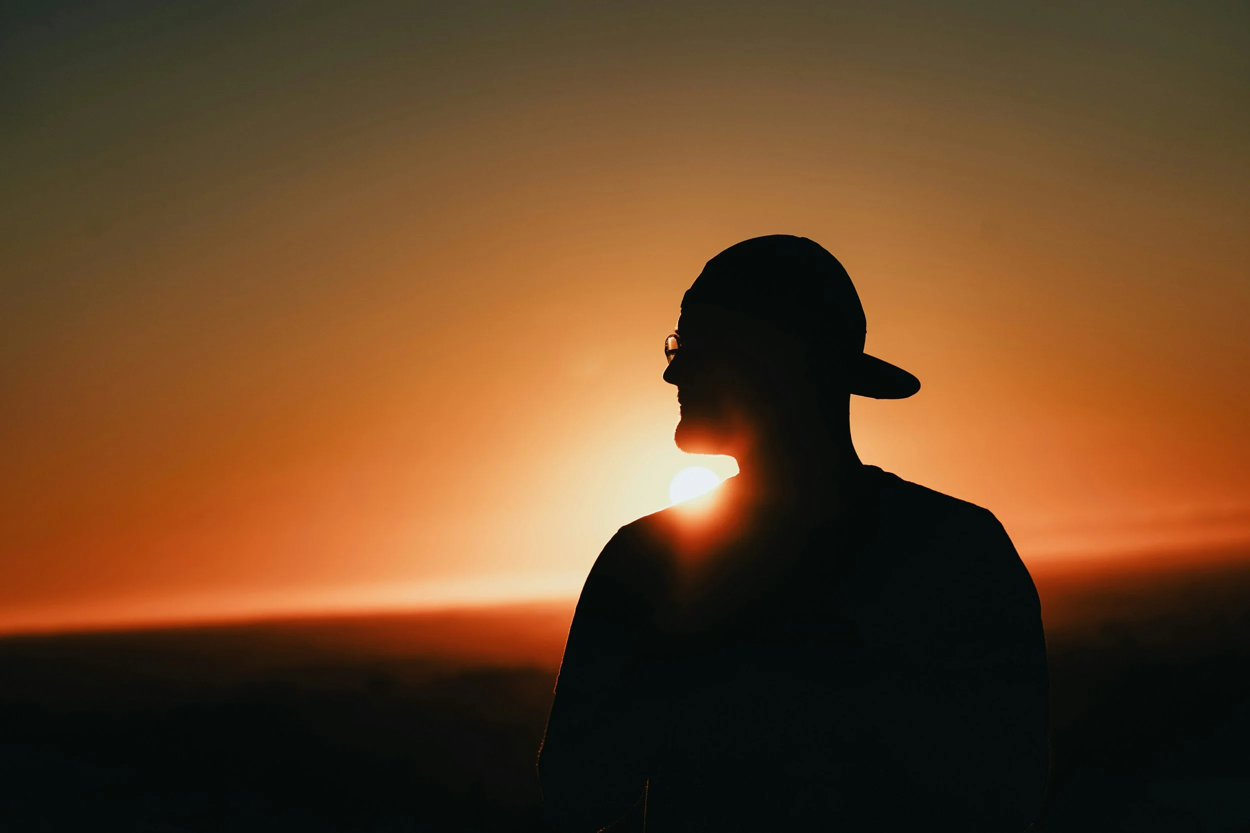 Silhouette of a person wearing a cap and glasses, standing in front of a sunset with the sun behind them.