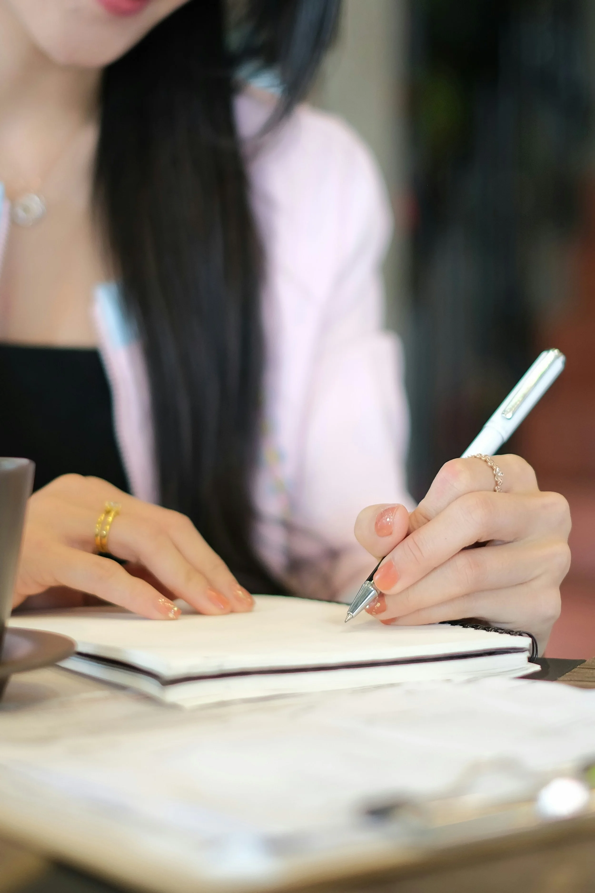 A woman writing in a notebook with a white pen, wearing rings and a necklace, with long black hair and a pink jacket.