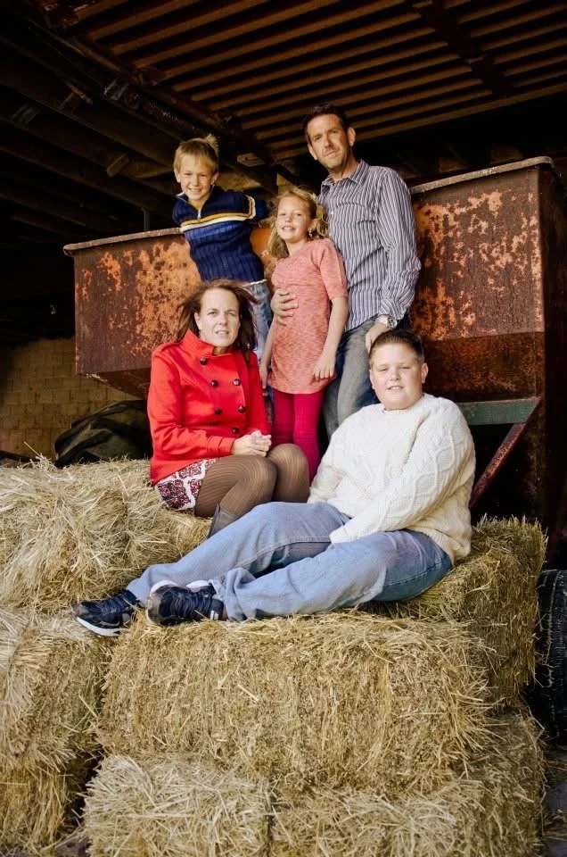 Marc Messinger and Family - Family picture with three children and two adults posing on hay bales inside a barn or stable. The children are sitting and standing, smiling at the camera.