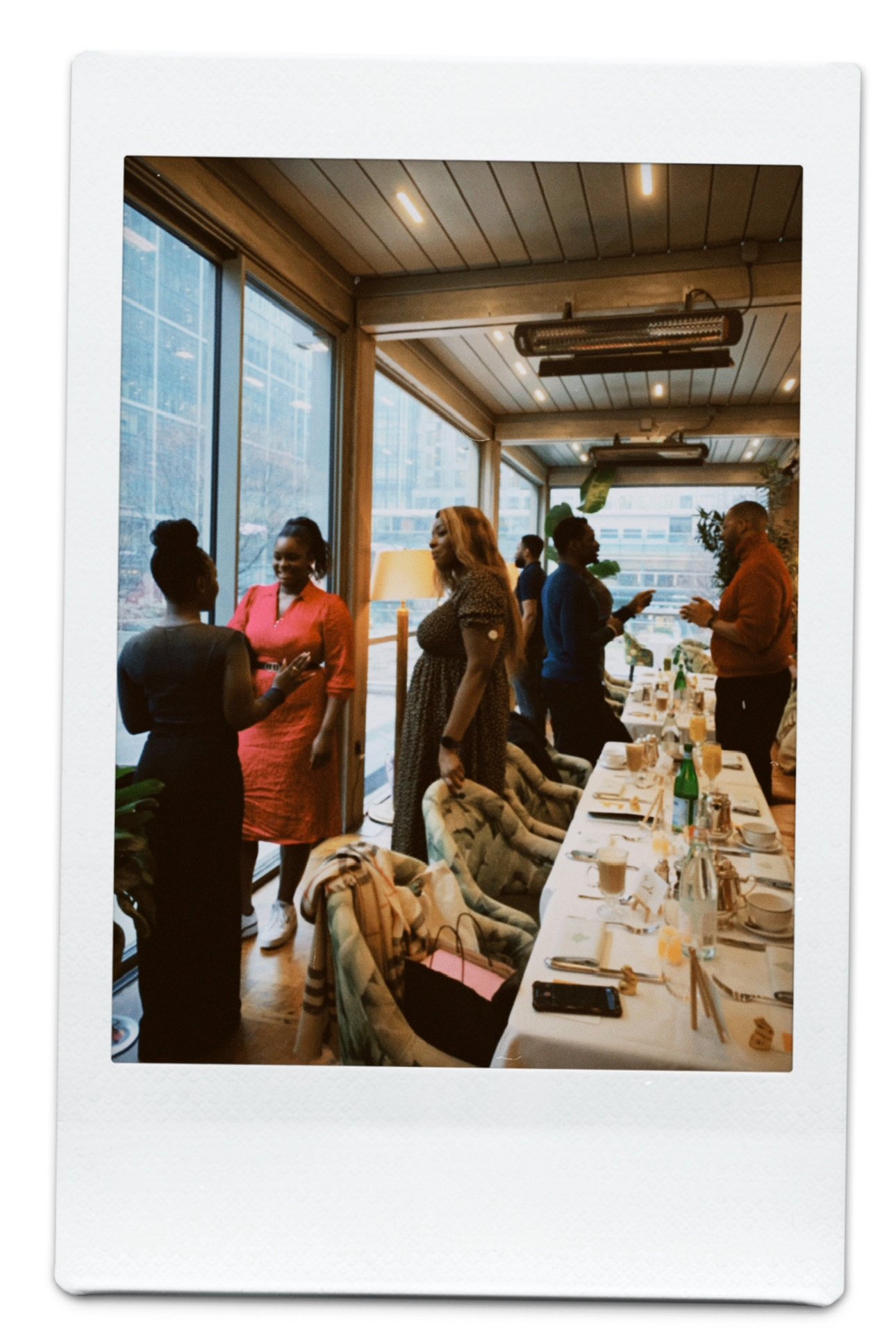The photo shows a group of women chatting and socializing in a well-lit restaurant with large windows, a long dining table set with drinks, plates, and utensils, and decorative plants.