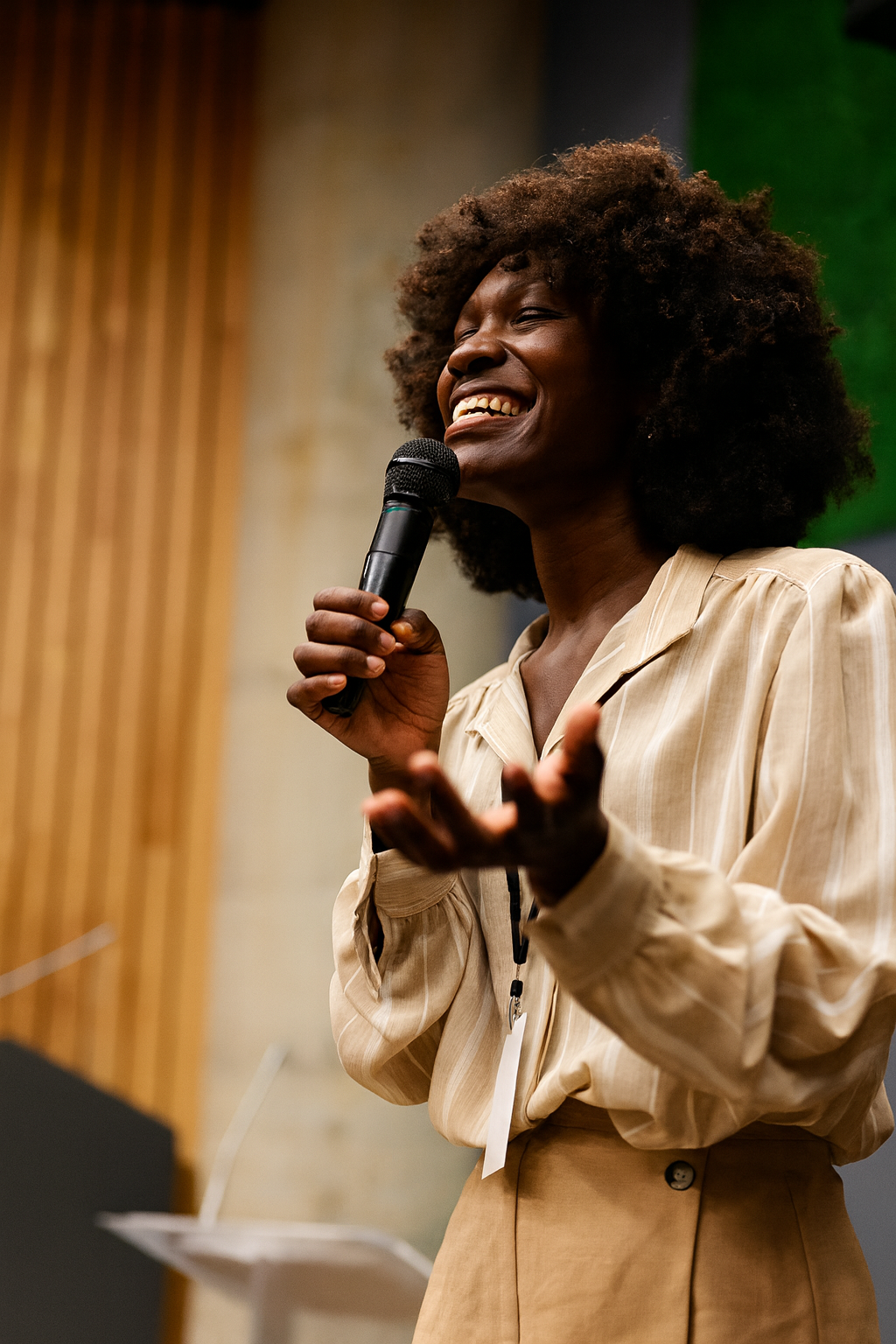A woman with natural curly hair smiling and speaking into a microphone during a presentation or speech.