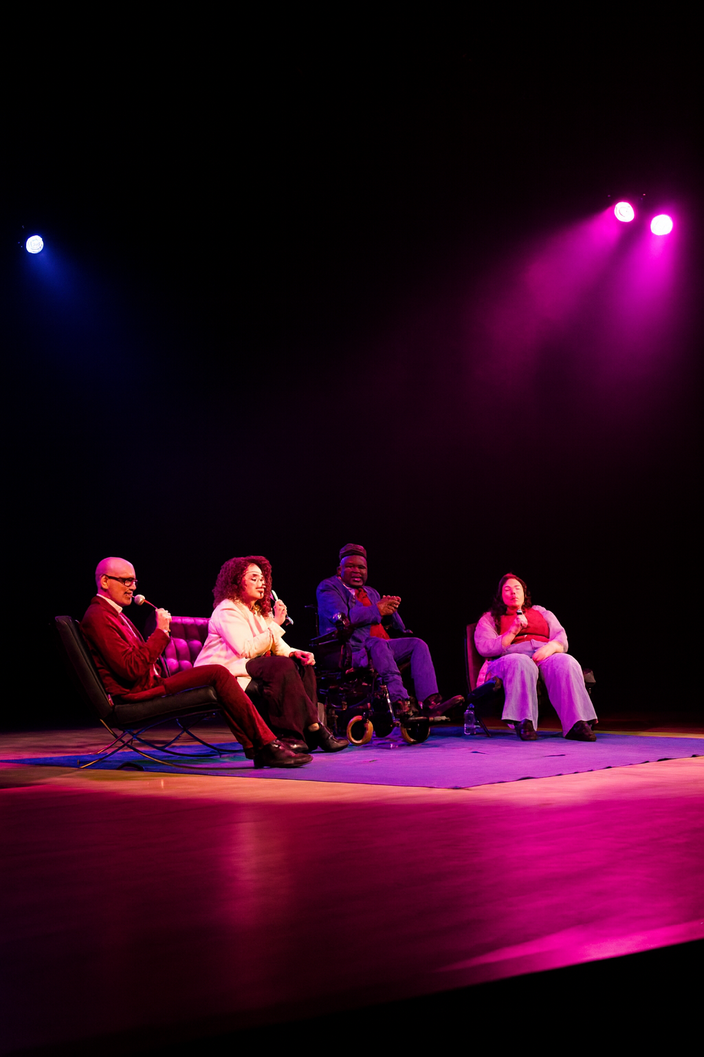 Four people seated on stage under colorful lights, participating in a discussion or panel, with microphones in their hands.