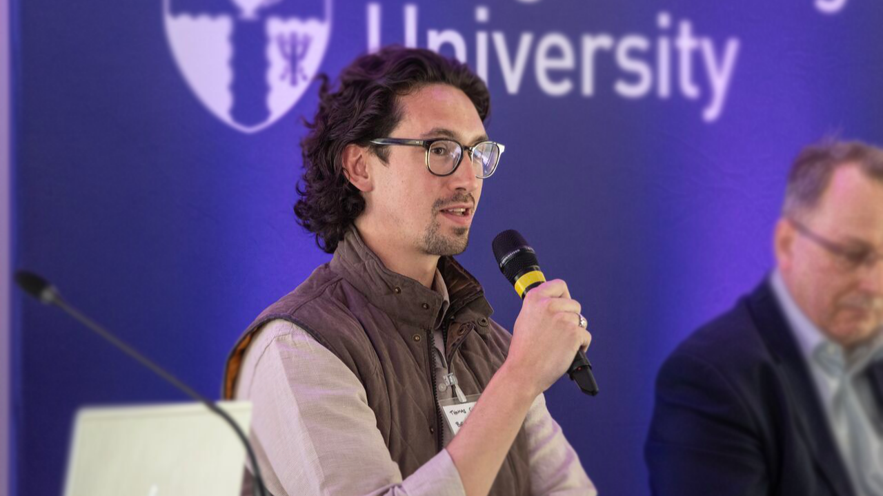 A man speaking into a microphone at a conference, with a university logo in the background.