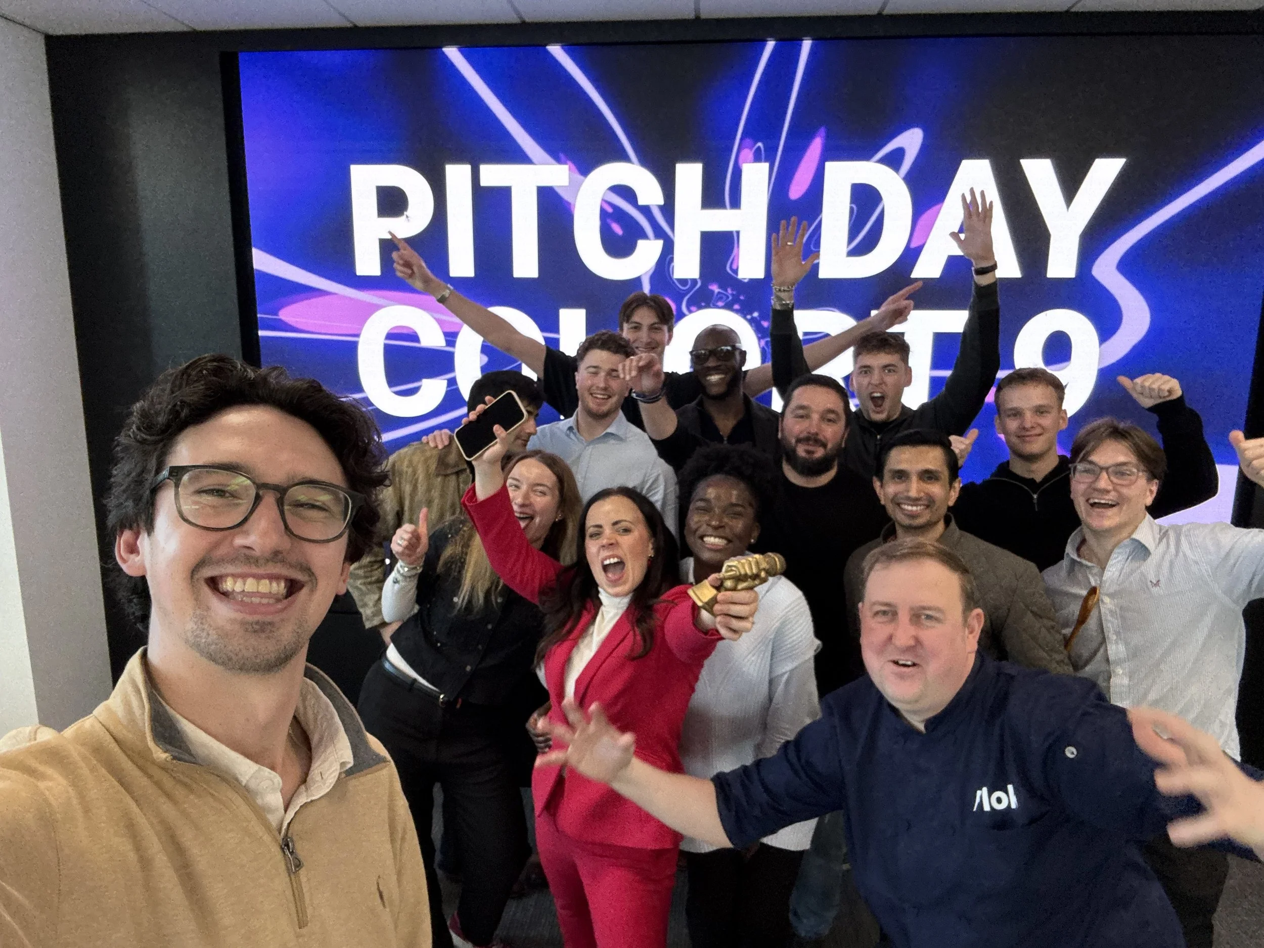 Group of diverse young people celebrating their victory at Pitch Day contest, taking a selfie with a large digital screen displaying 'Pitch Day Contest' in the background.