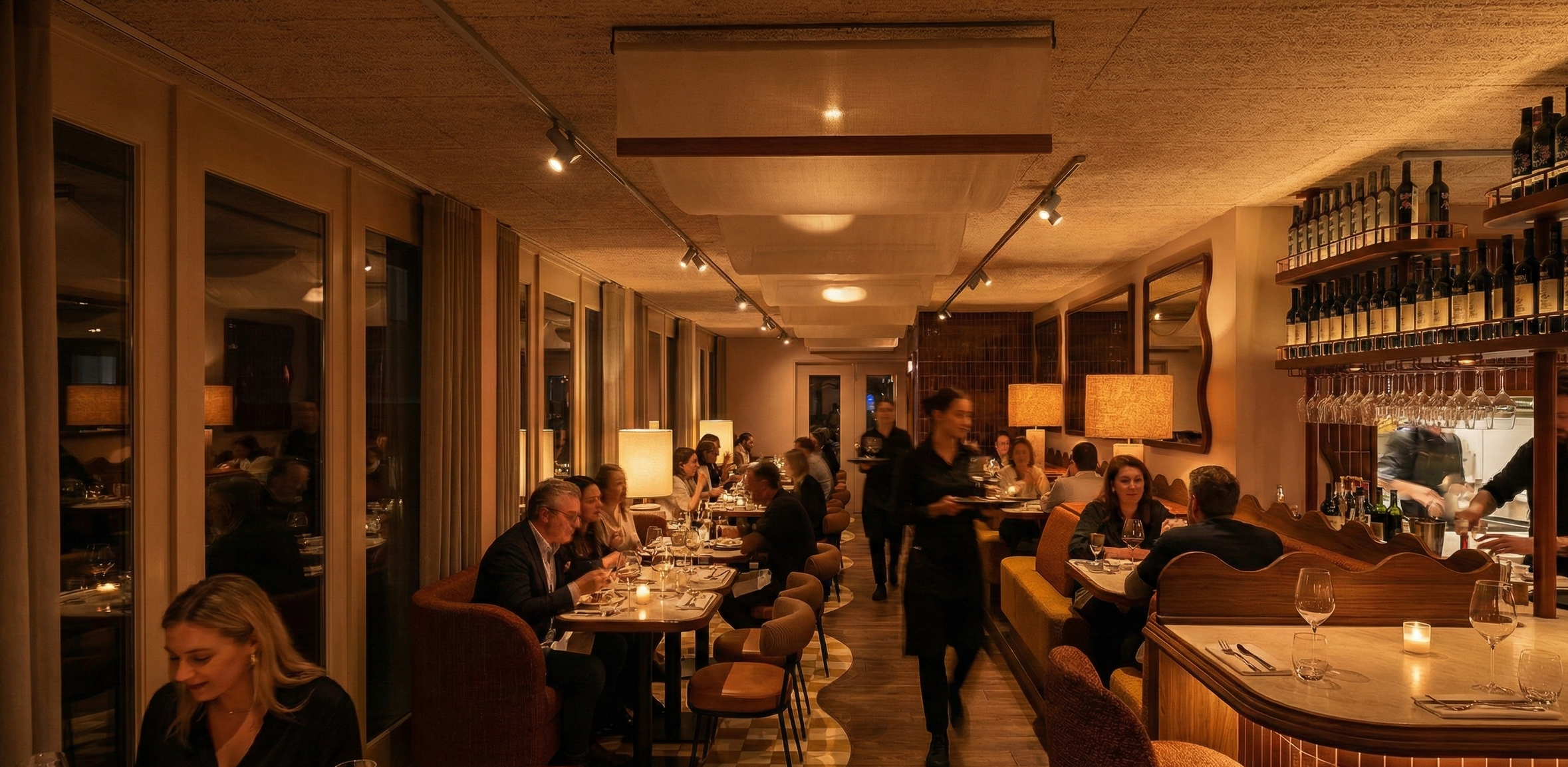 Een sfeervol restaurant met gasten die genieten van hun maaltijd, terwijl obers serveren. Warme verlichting, grote ramen en houten meubels creëren een gezellige ambiance.