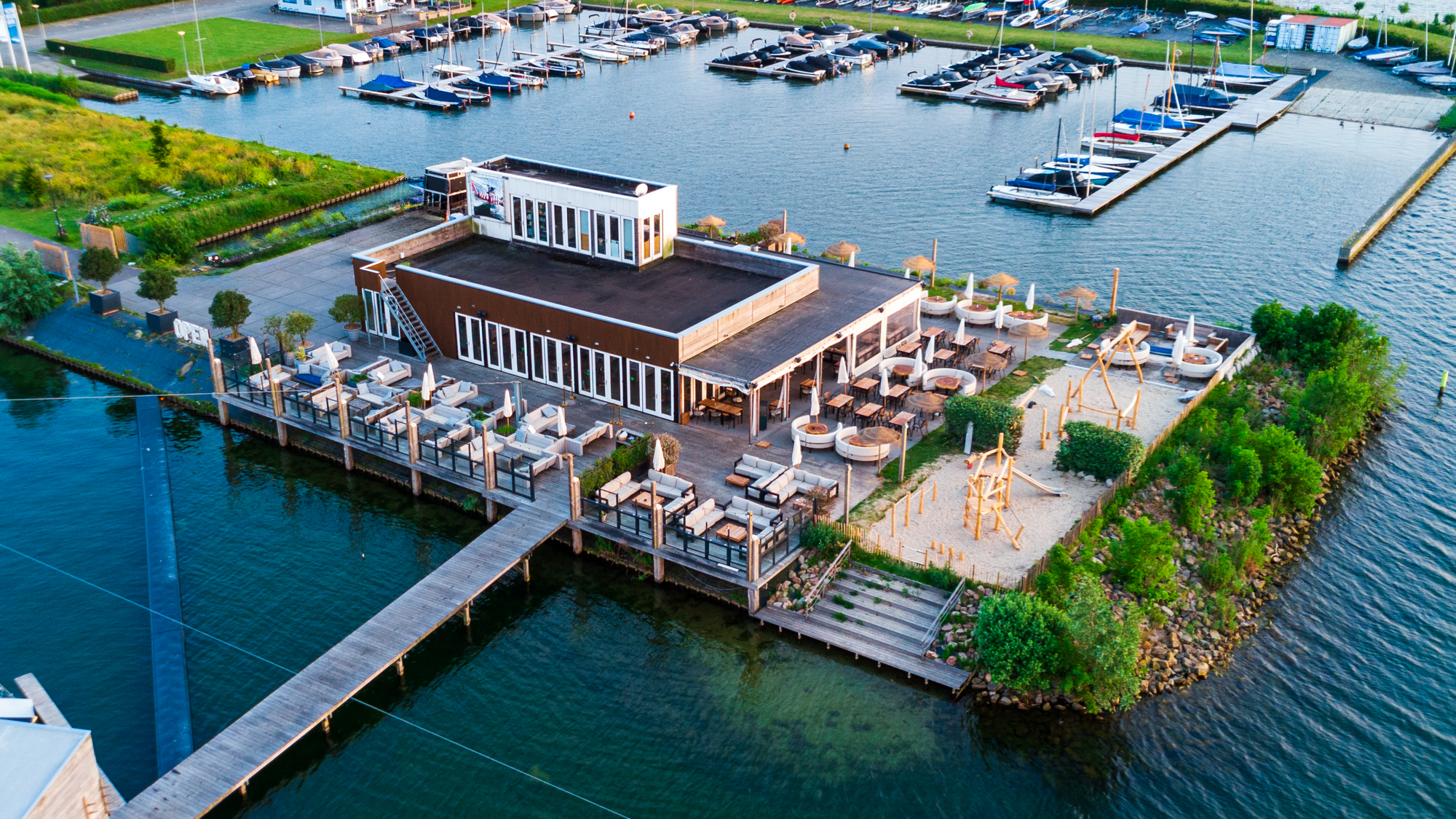 Luchtfoto van een modern restaurant met een terras aan het water, naast een jachthaven met boten.