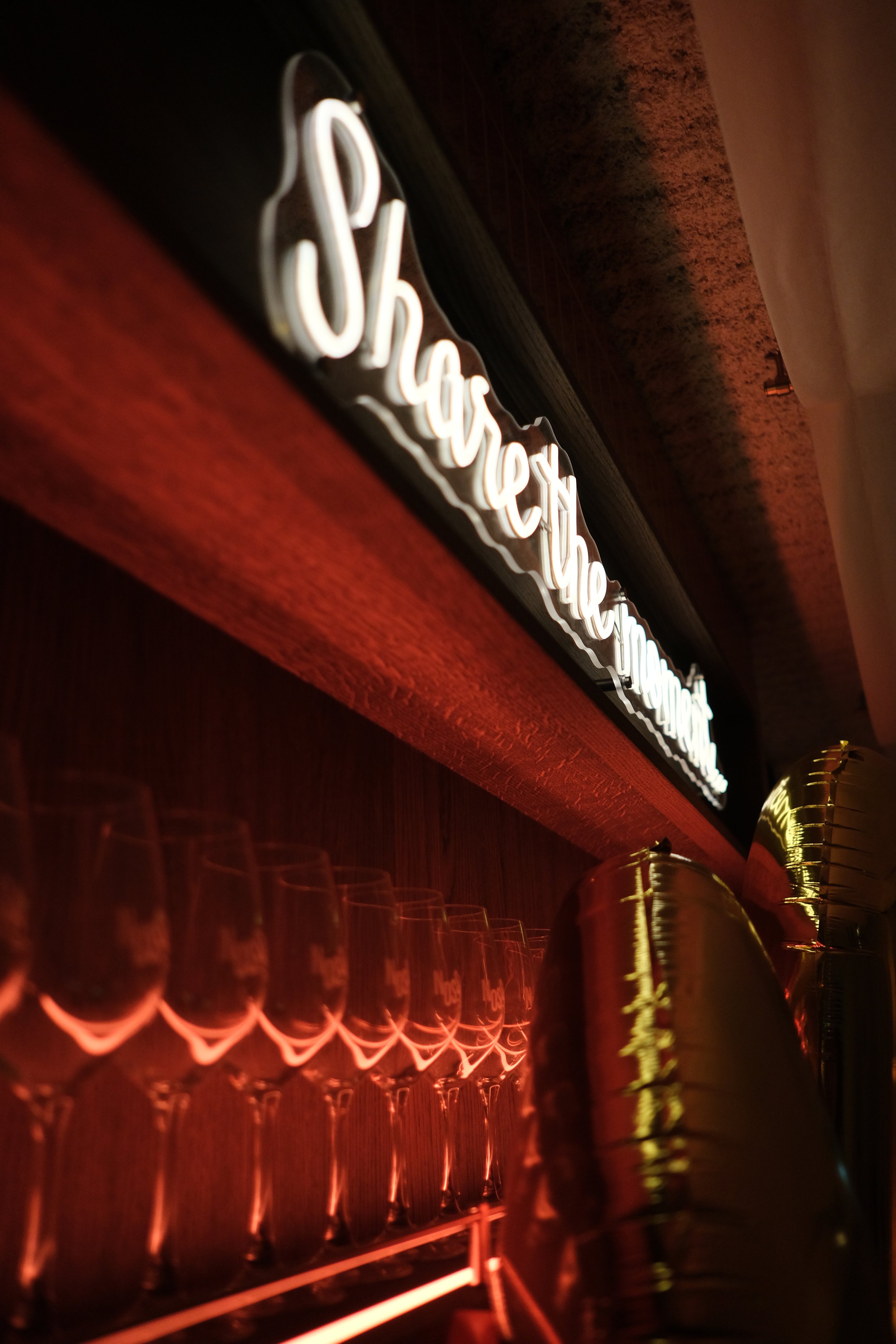 Neonbord met de tekst 'Share the moment' boven een rij lege wijn- of champagneglazen in een bar met warme rode verlichting.