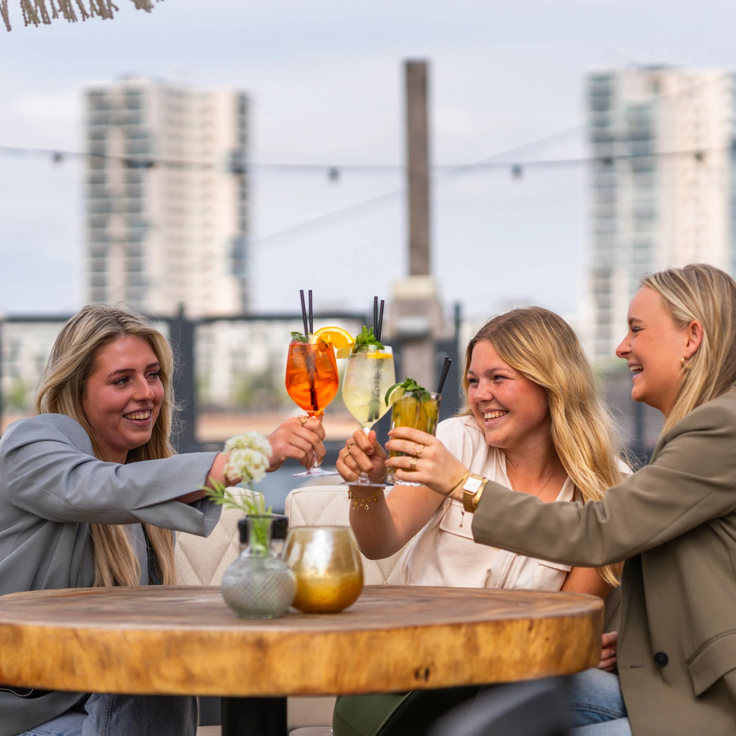 Vier vrouwen lachen en proosten met kleurrijke cocktails op een terras met uitzicht op hoge gebouwen.