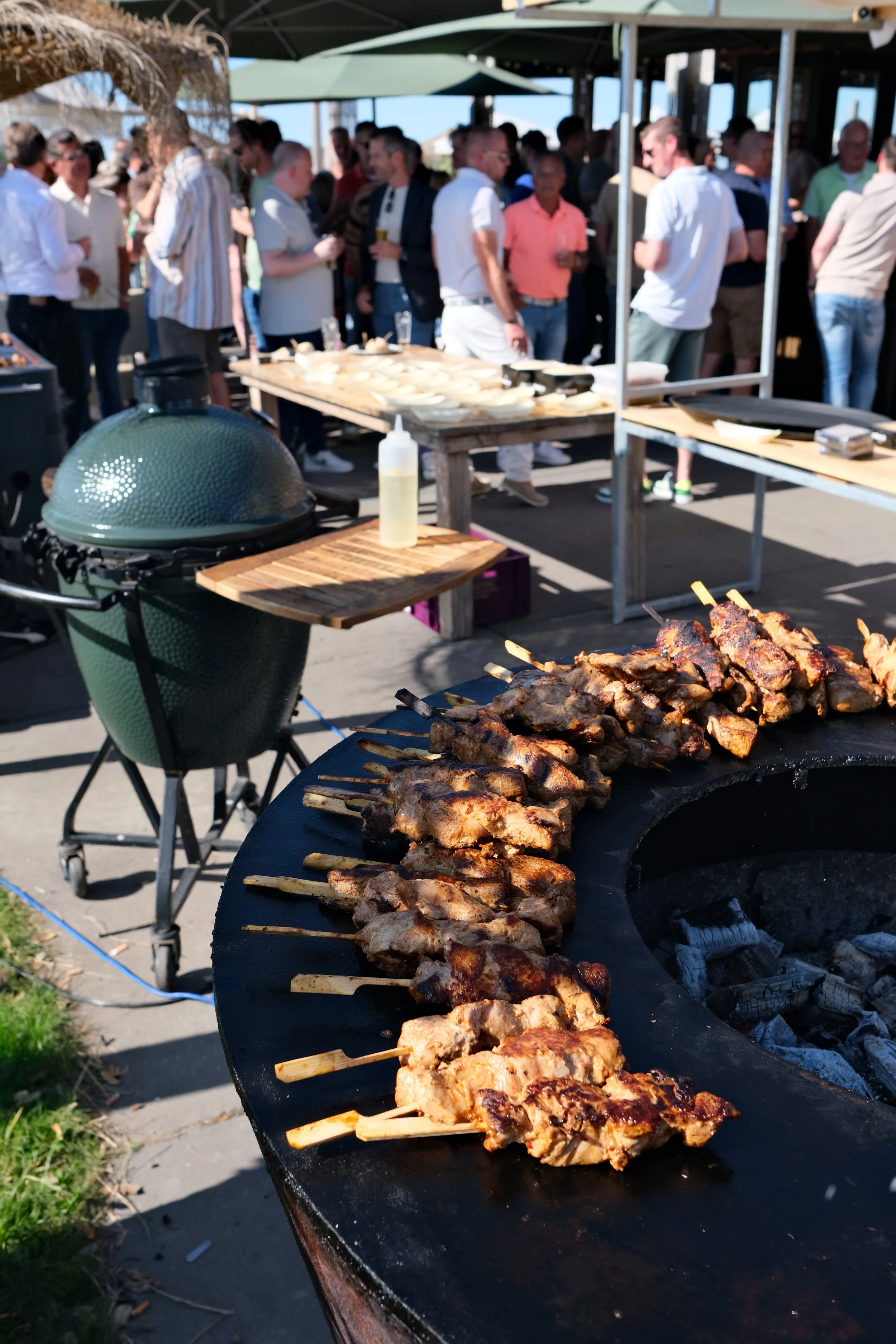 Gesuikerde BBQ-spiezen met vlees op de barbecue, omgeven door mensen die genieten van een buitenfeest.