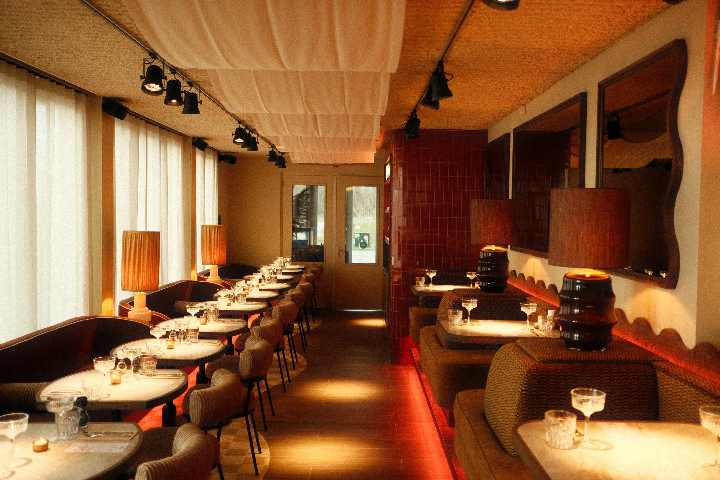Een gezellig restaurant met warme verlichting, tafels gedekt met glazen en bestek, en comfortabele stoelen. Grote witte gordijnen bedekken de ramen en er is een houten vloer en muur met rode tegels.