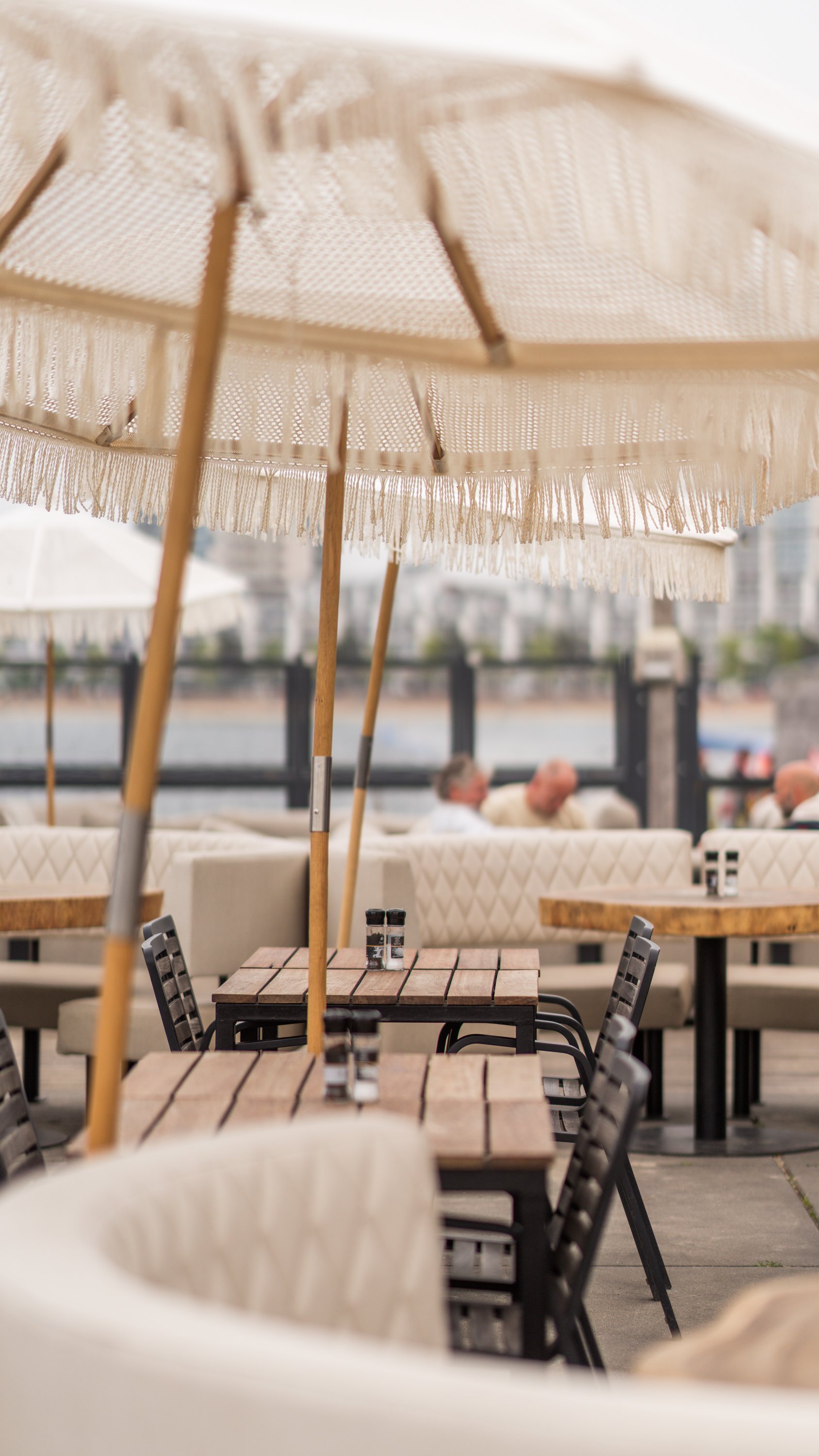 Een terras met houten tafels, zwarte stoelen en grote parasols met fringes, waar mensen in de achtergrond aan tafel zitten.