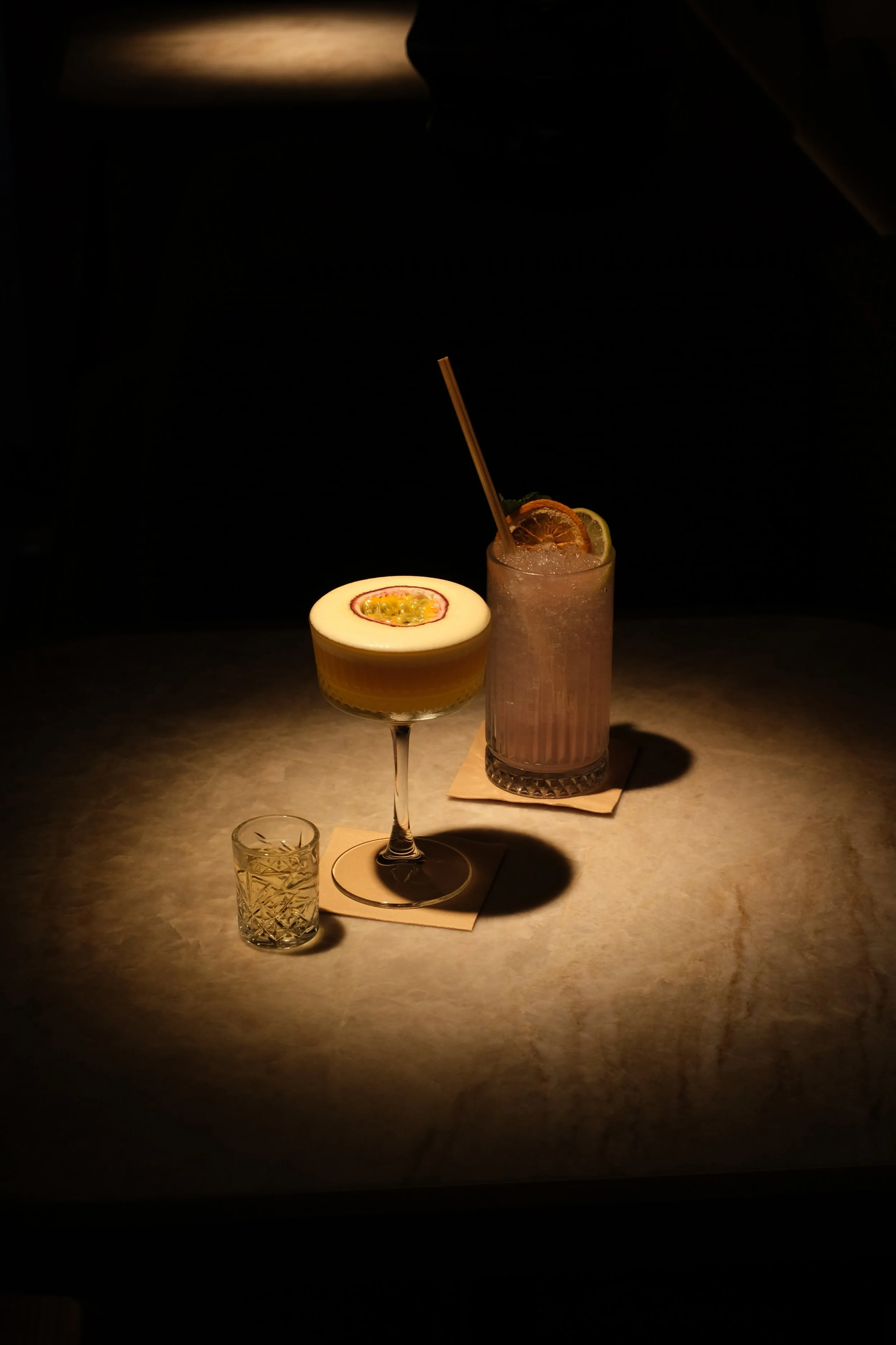 Drie verschillende cocktails op een tafel, met een spotlight erop, met donkere achtergrond. Eén cocktail met een passievrucht en één met citroen, en een klein glas erbij.