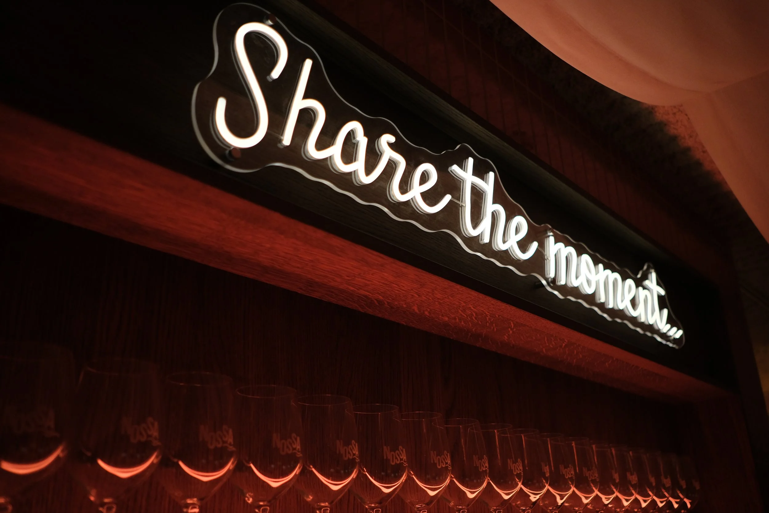 Neonbord met de tekst 'Share the moment' boven een rij wijn- of champagneglazen in een bar of restaurant.