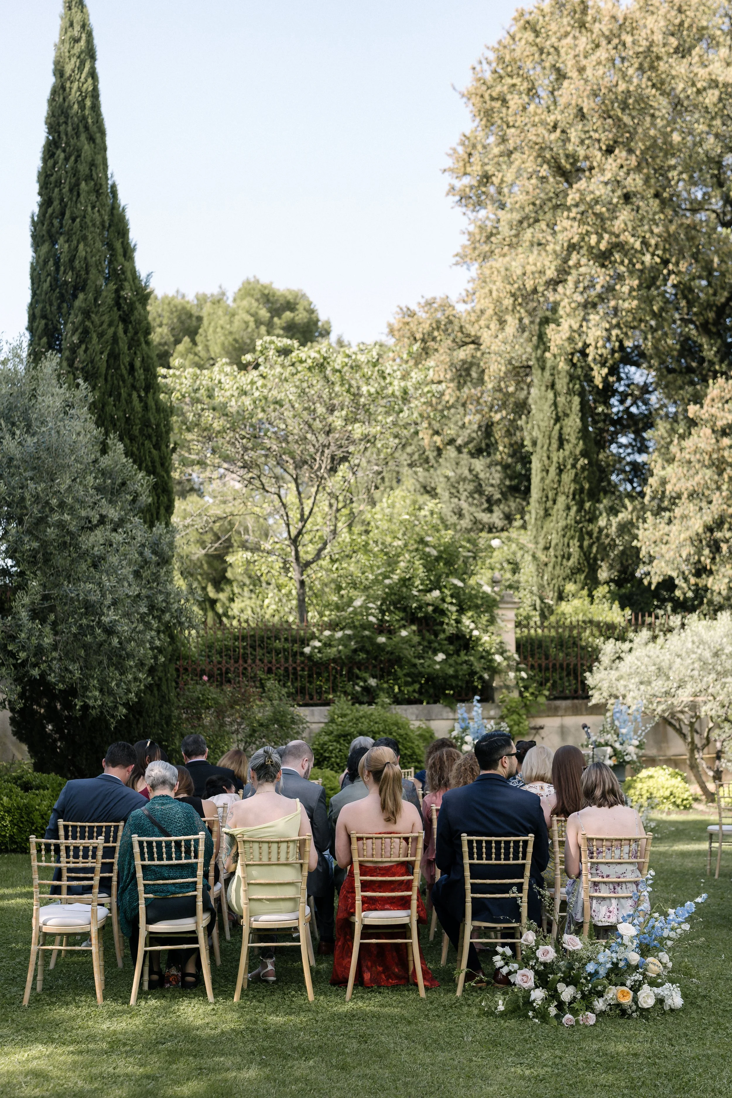 Groupe de personnes assises dans le jardin d'un domaine provençal lors d'une cérémonie laïque en plein air, entouré d'arbres et de fleurs, avec des chaises en bois et un arrangement floral à l'avant.