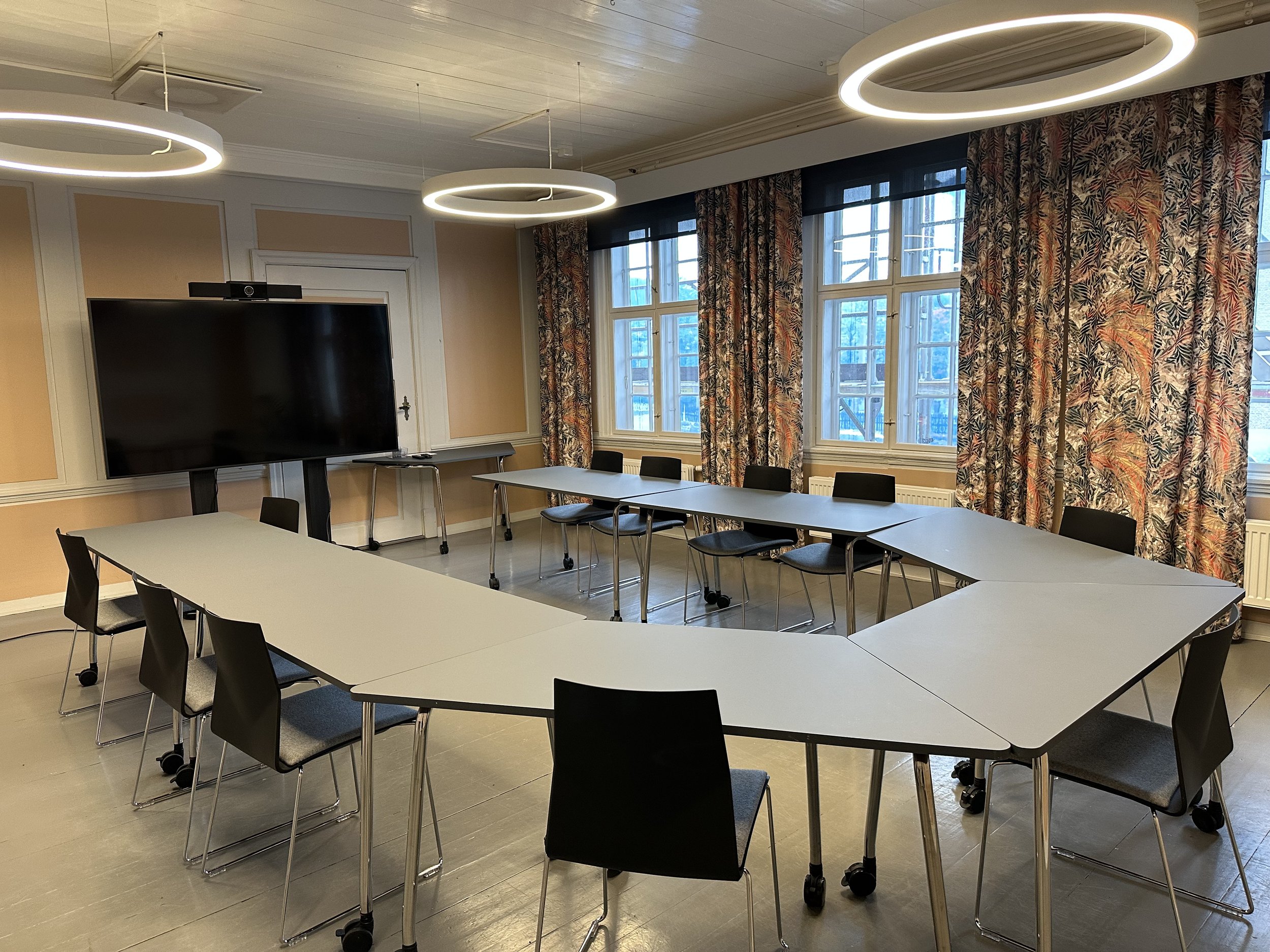 Et tomt møterom med bord plassert i hestesko-formasjon, og svarte stoler rundt. Det er en stor TV på stativ ved den ene veggen, blomstrete gardiner og tre moderne sirkulære lamper i taket.