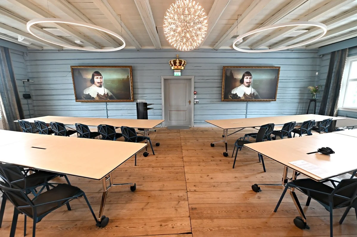 Auditoriesalen med bord og stoler plassert i forelesnings-formasjon. Blå tømmervegger og to TV-er på veggen.