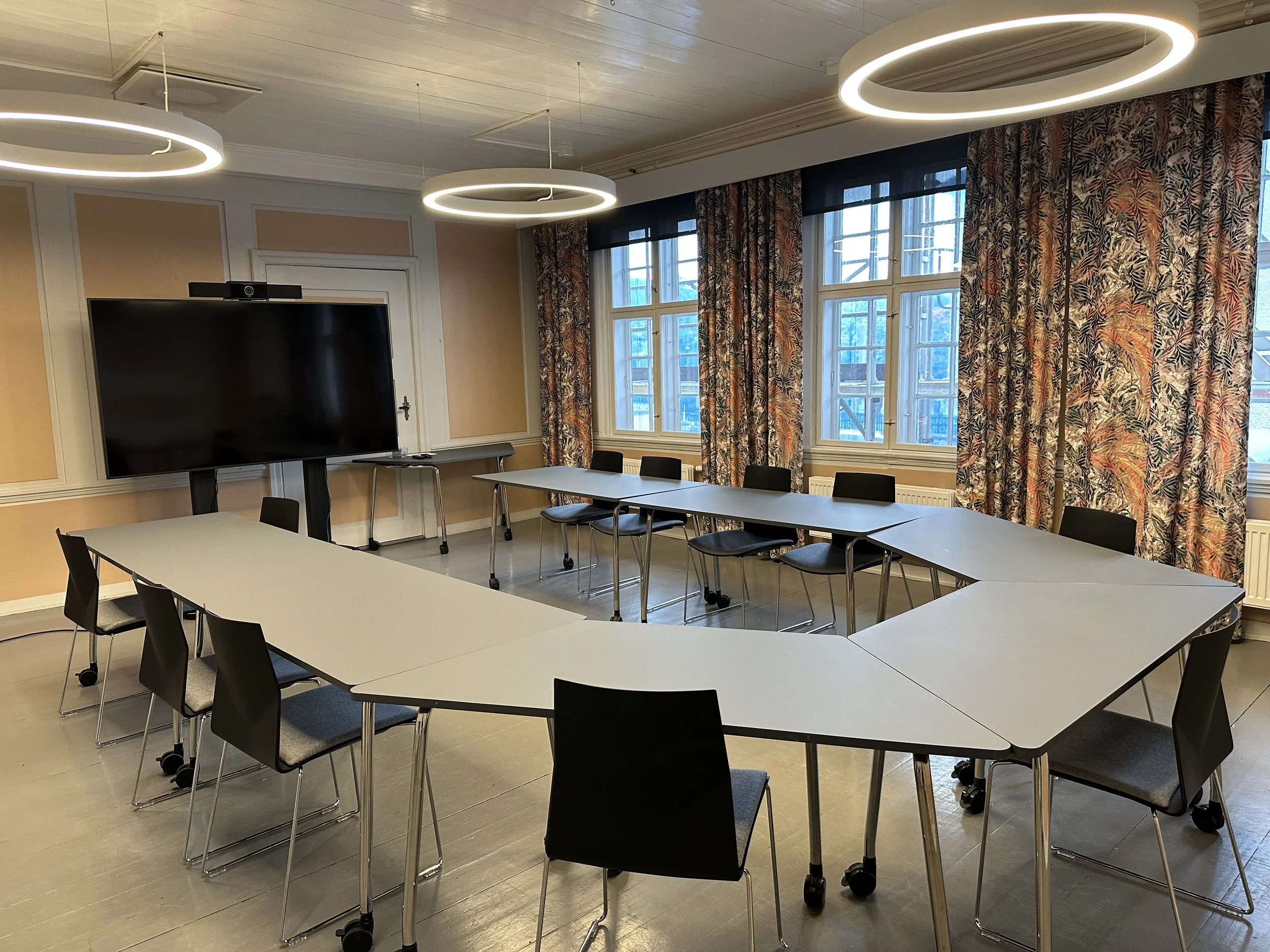 Et tomt møterom med bord plassert i hestesko-formasjon, og svarte stoler rundt. Det er en stor TV på stativ ved den ene veggen, blomstrete gardiner og tre moderne sirkulære lamper i taket.