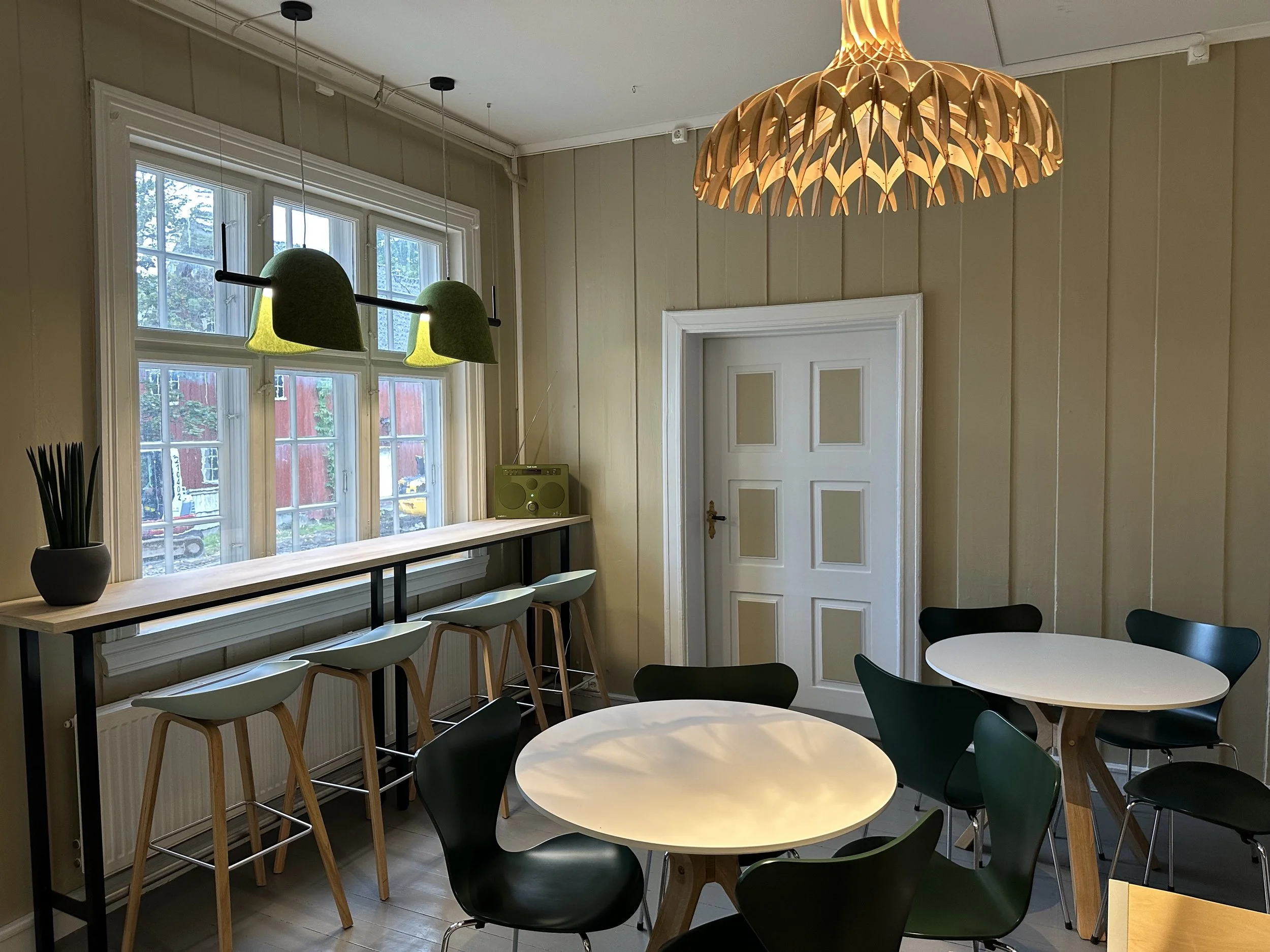 Kaféen i Bergseminaret med runde bord, varm belysning, gule vegger, hvit dør og grønne lamper over et høyt bord med tilhørende barstoler ved et vindu.