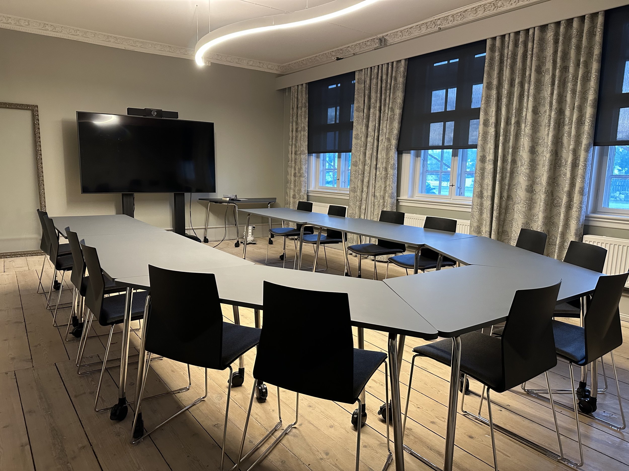 Et tomt møterom med bord plassert i hestesko-formasjon, og svarte stoler rundt. Det er en stor TV på stativ ved den ene veggen.
