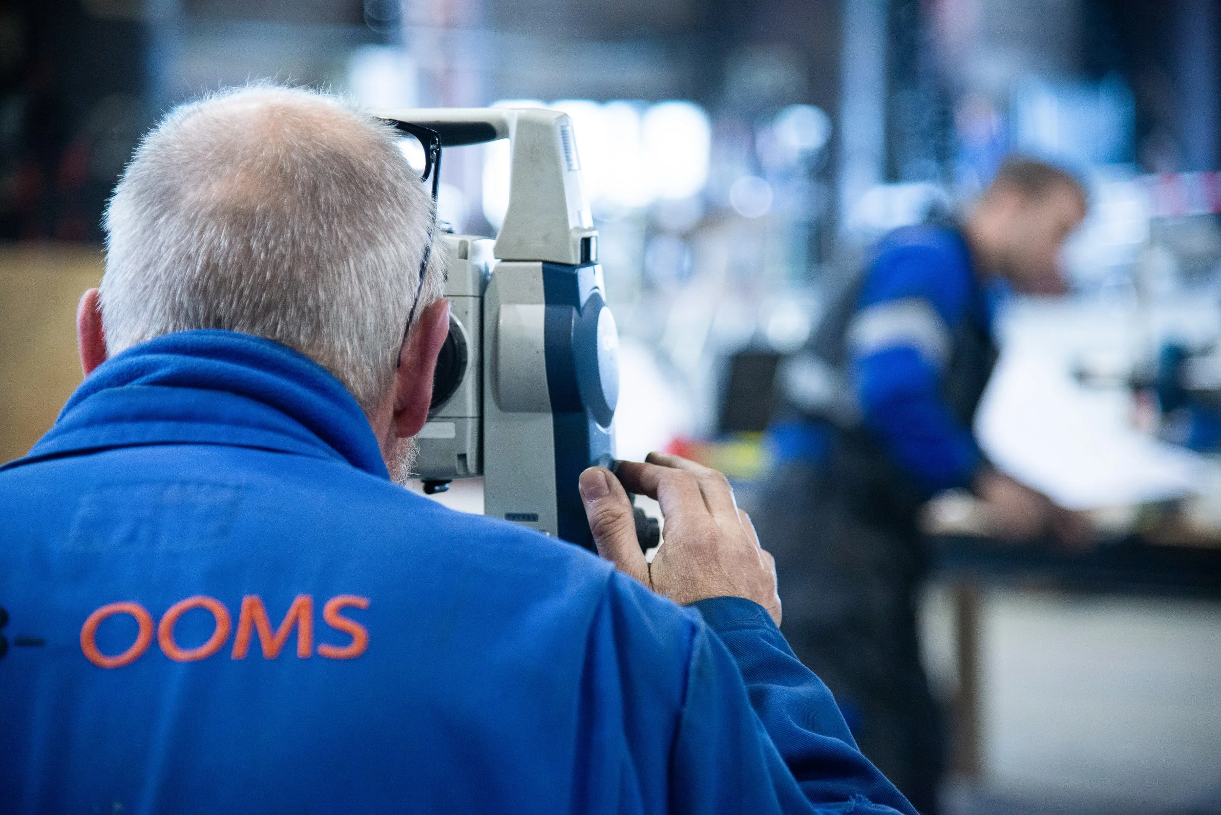 A man with gray hair and glasses using a machine in a workshop, with a woman working at a table in the background.