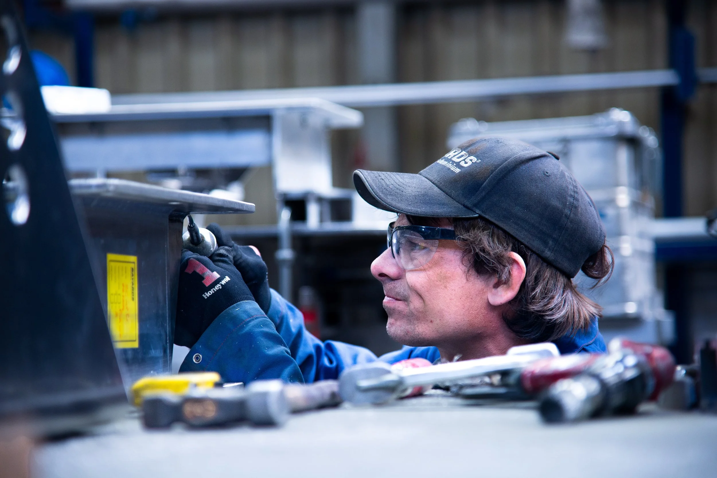 A man wearing safety glasses, a black cap, and gloves is working on a metal structure with tools around him in an industrial workshop.