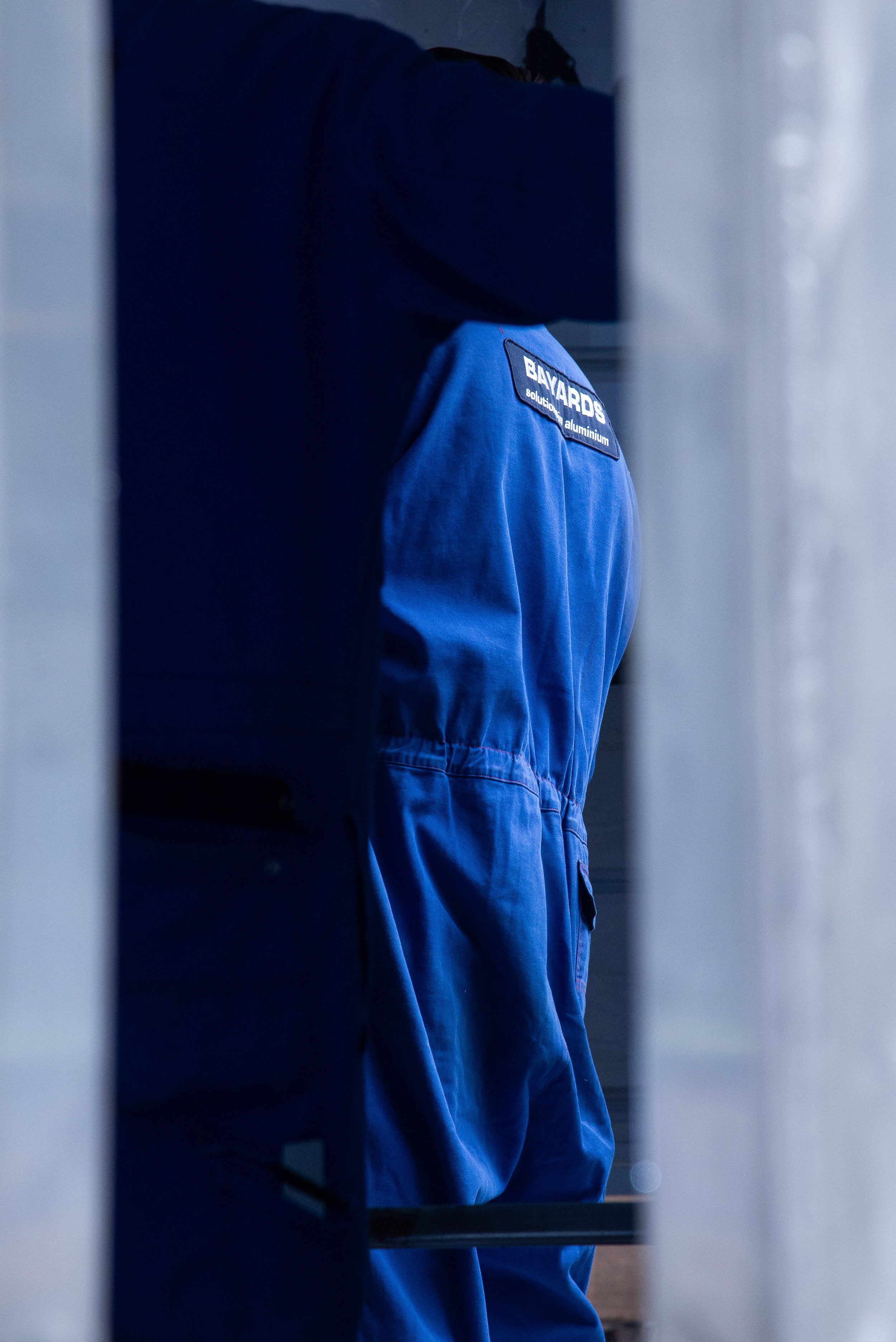 Close-up view of a person wearing a blue jumpsuit with a Bayards logo patch on the shoulder, peeking out from behind a curtain.
