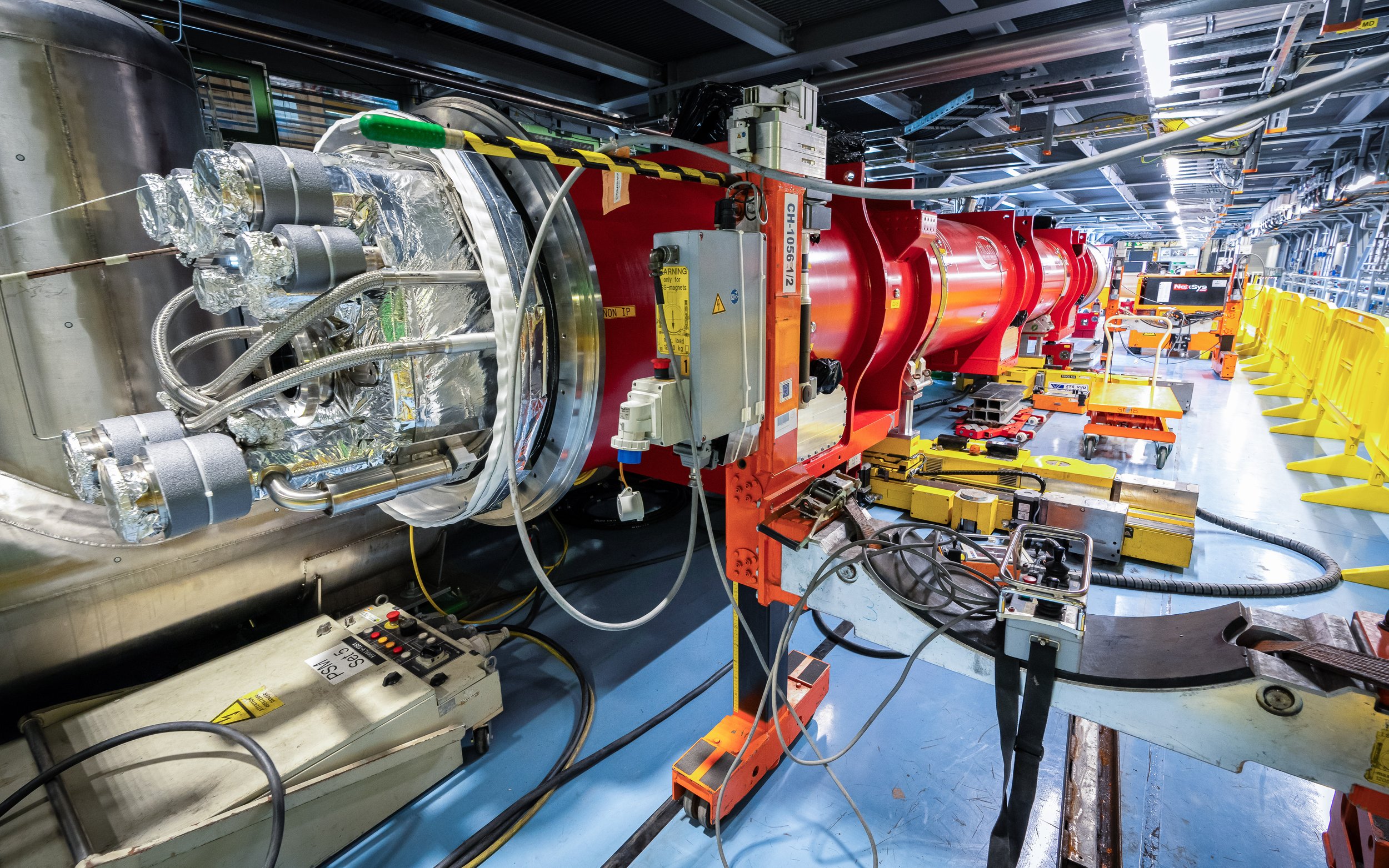 Large industrial equipment with a red cylindrical component, connected to various wires, tubes, and insulation material, situated in a factory setting with yellow safety barriers and tools nearby.