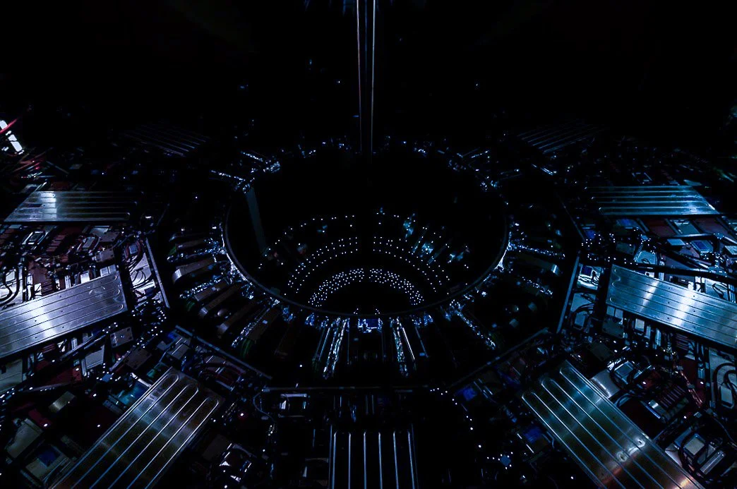 Inside a large particle laser collider with concentric rings of blue lights and metallic components, viewed from above in a dark environment.
