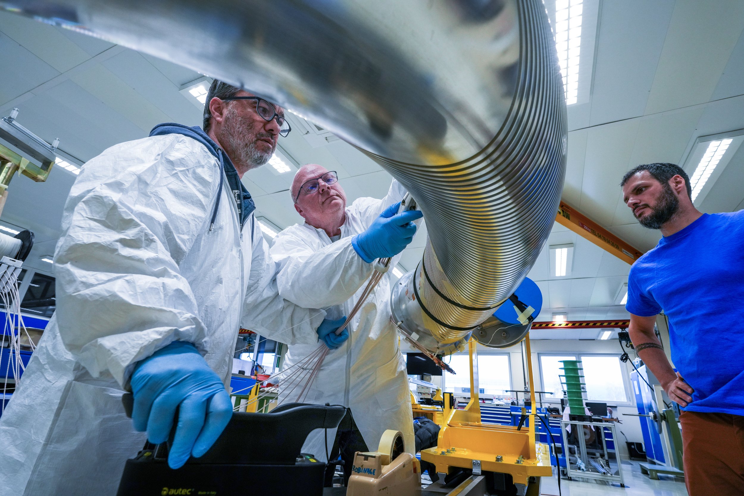 Three scientists or engineers in lab coats and gloves working on a large metallic pipe or tube in a high-tech laboratory or research facility.