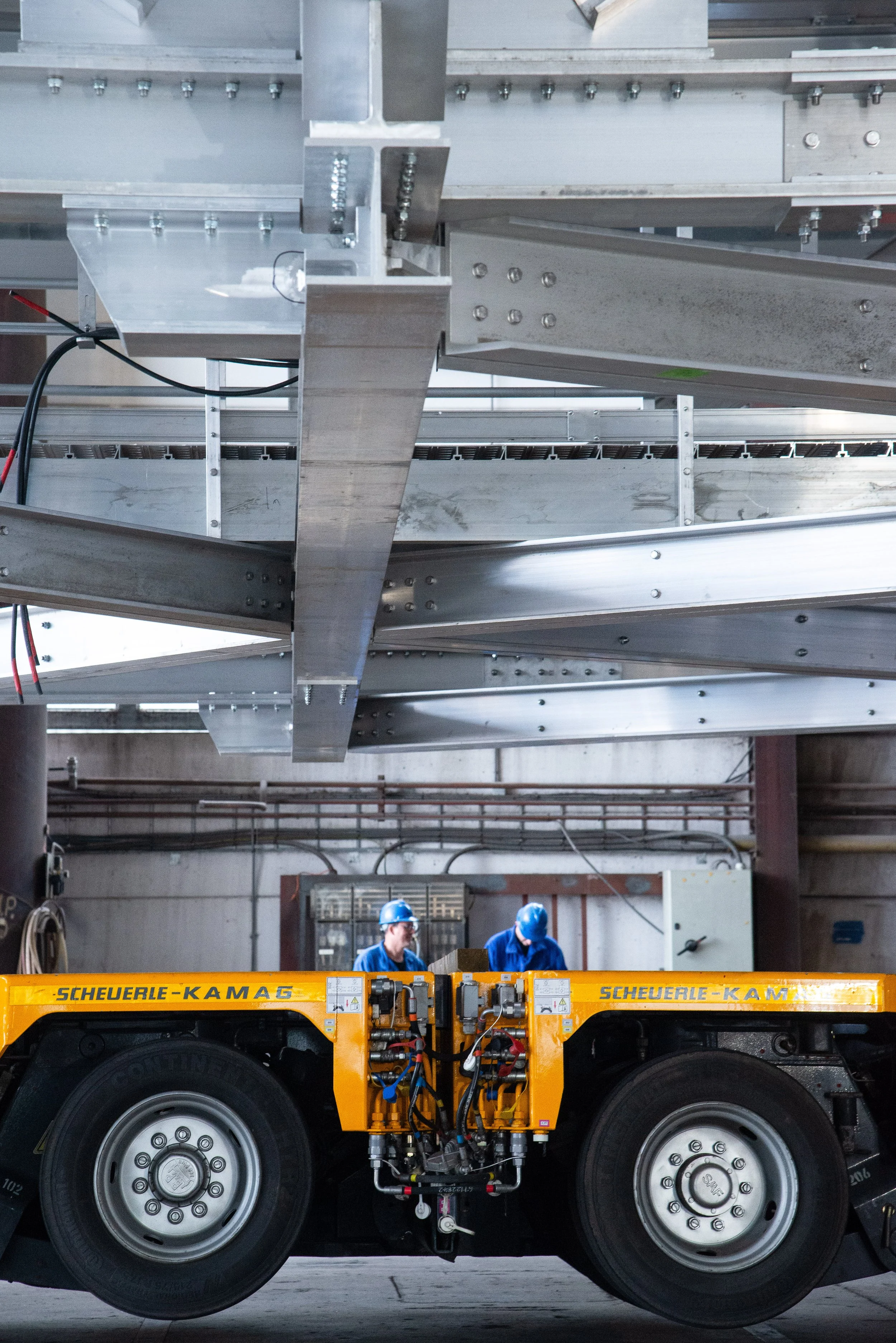 Workers in blue uniforms and helmets working with industrial machinery in a factory setting. The machinery includes large metal beams and a yellow hydraulic lift.