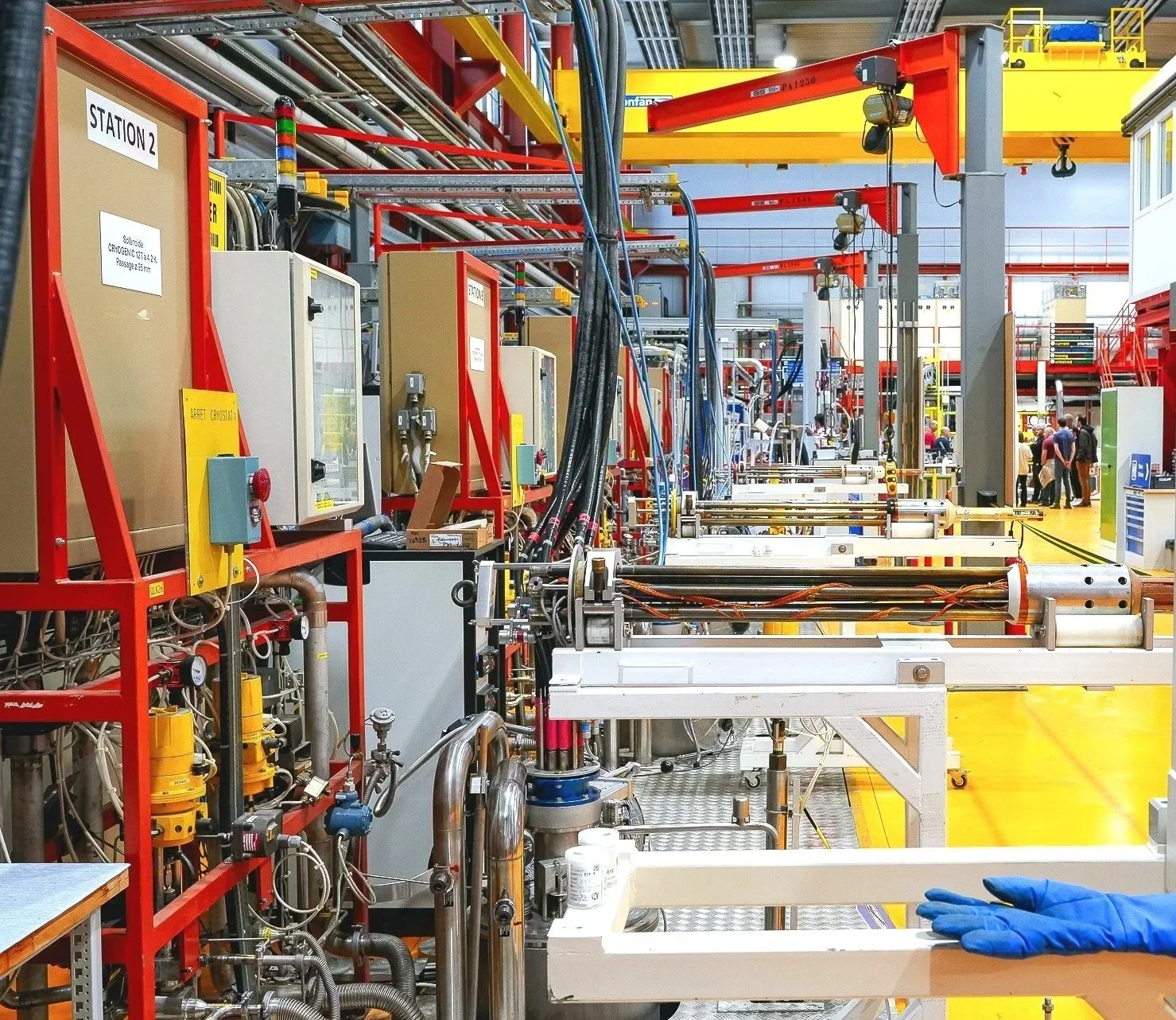 Laboratory or industrial facility with machinery, control panels, pipes, and a person wearing blue gloves in the foreground.