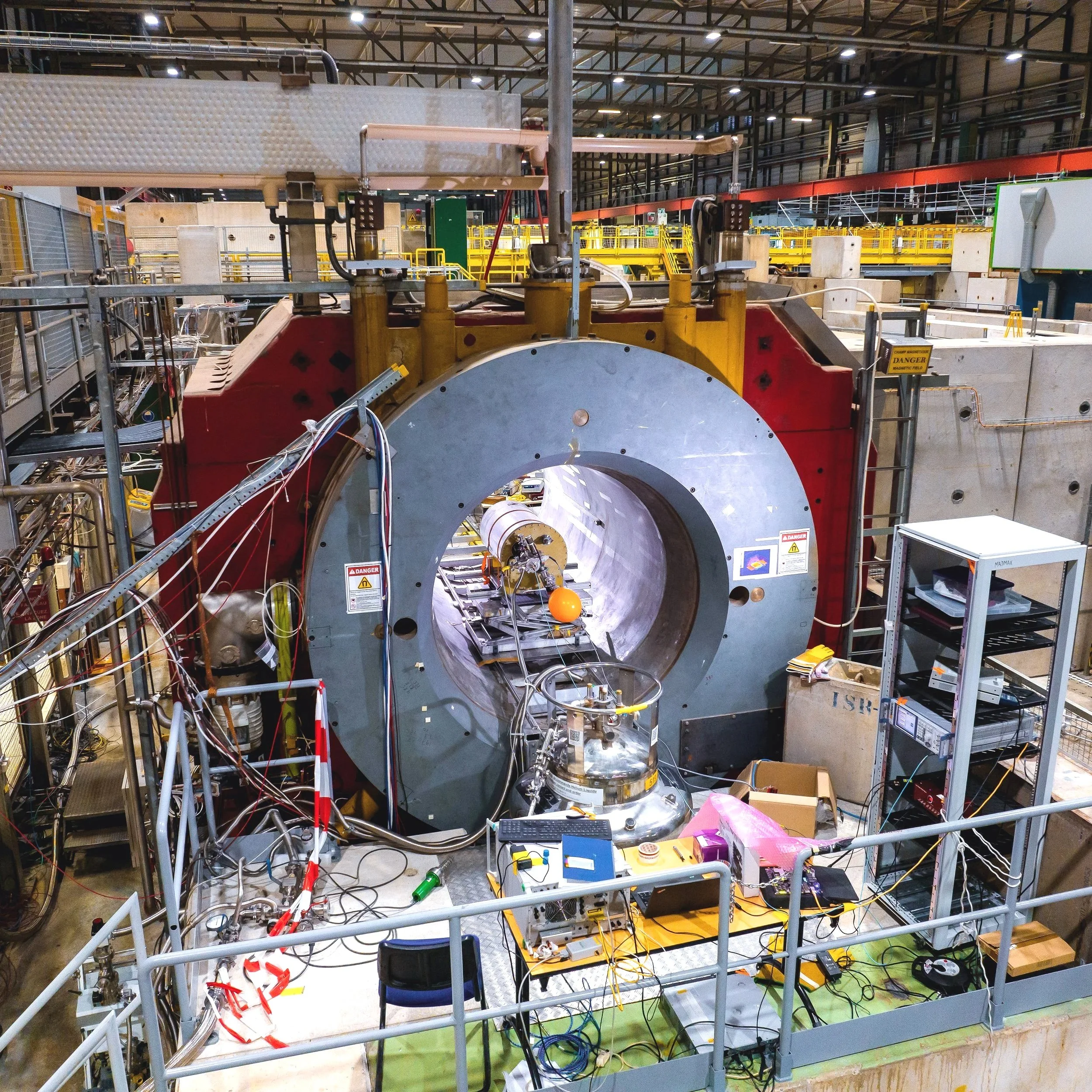 A large particle accelerator or collider in a high-tech laboratory with various equipment, wires, and safety signs.