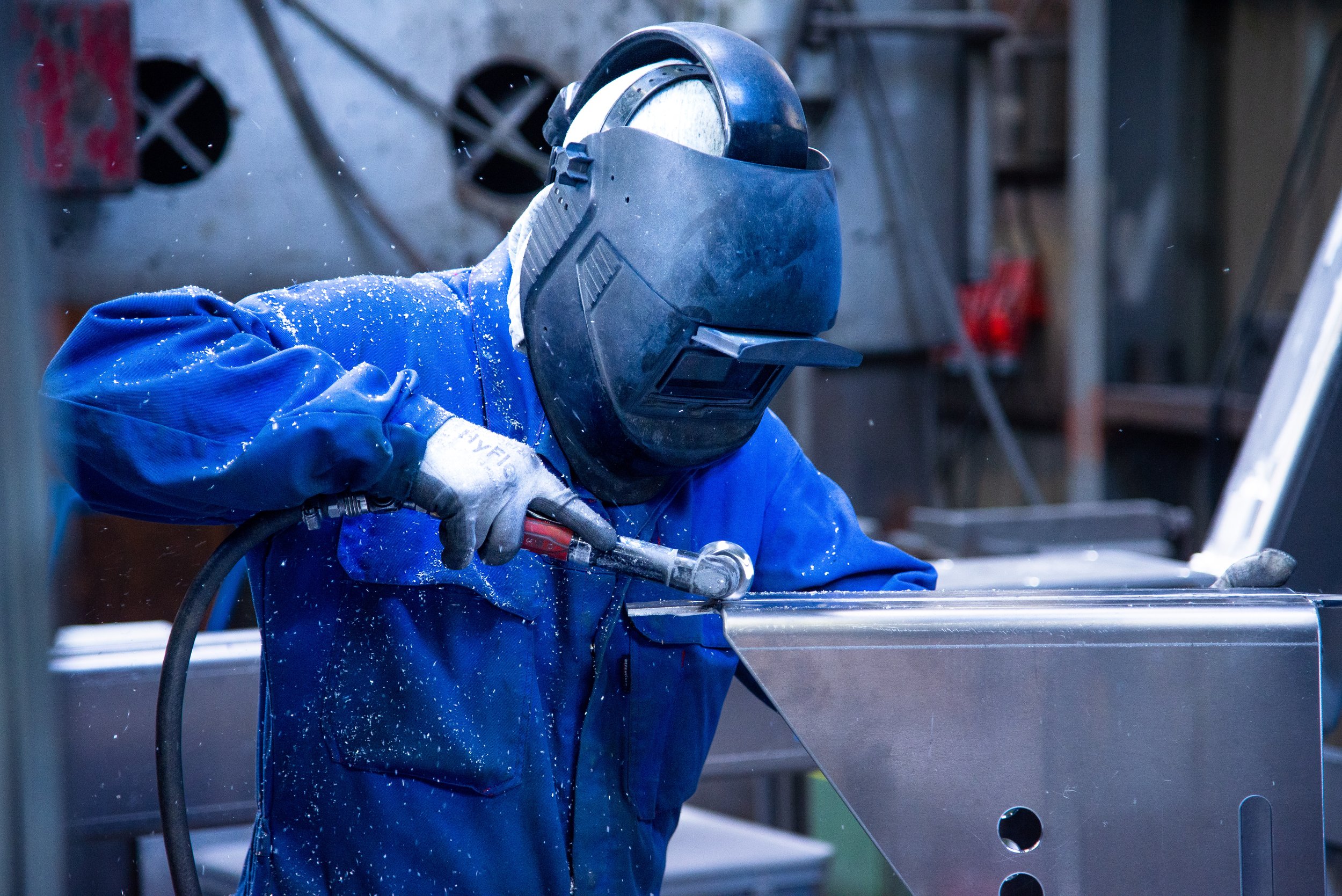 A person wearing a welding helmet and blue work jacket welding a metal piece in a workshop.