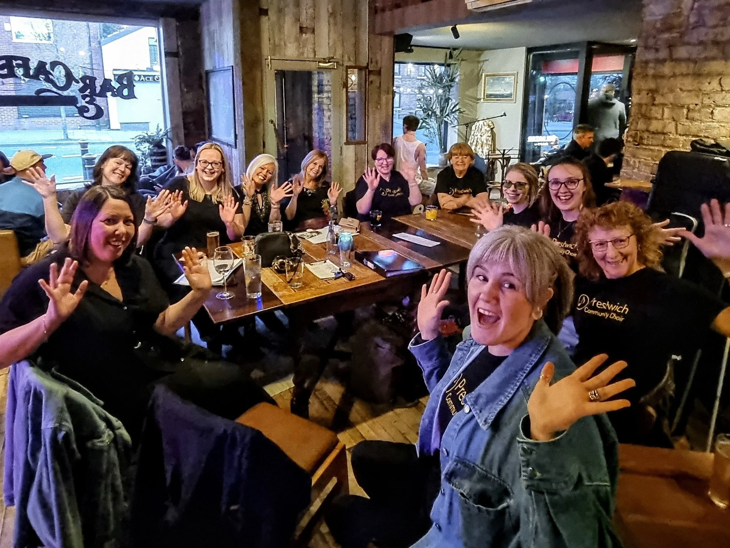 Group of women smiling and waving at a dining table in a restaurant or pub, with drinks and menus on the table, some wearing black shirts with yellow text.