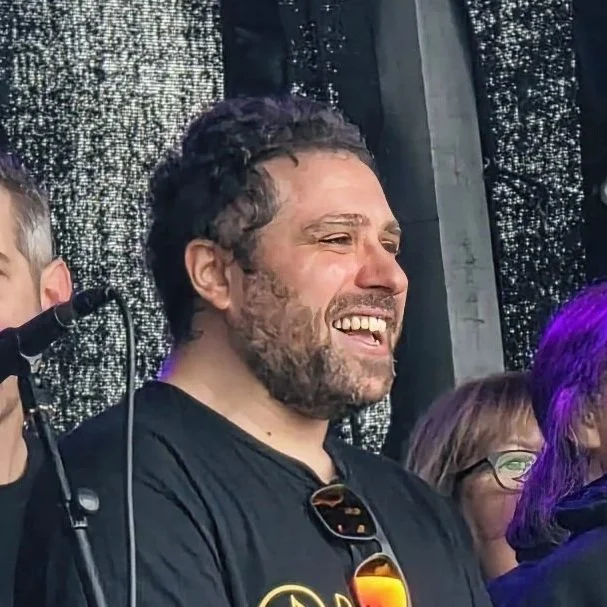 A man crying and smiling at an outdoor event, wearing a black shirt with sunglasses hanging from his collar, with a microphone in front of him and a glittery backdrop.