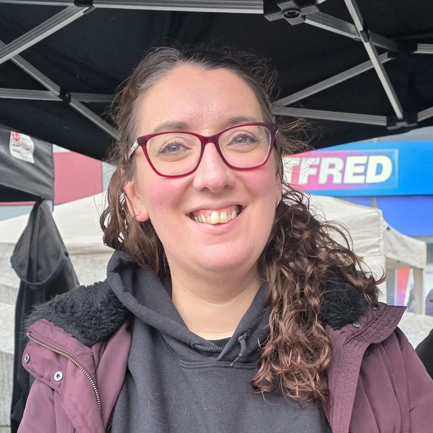 A woman with curly brown hair and red glasses smiling at the camera, standing outdoors under a black canopy with a red, white, and blue sign in the background.